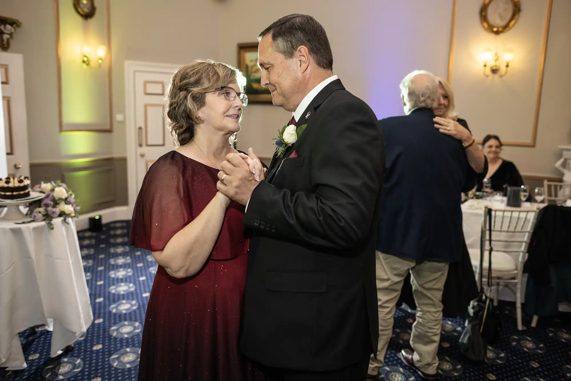 A man in a suit and a woman in a maroon dress dance together in a decorated room, while another couple dances in the background.