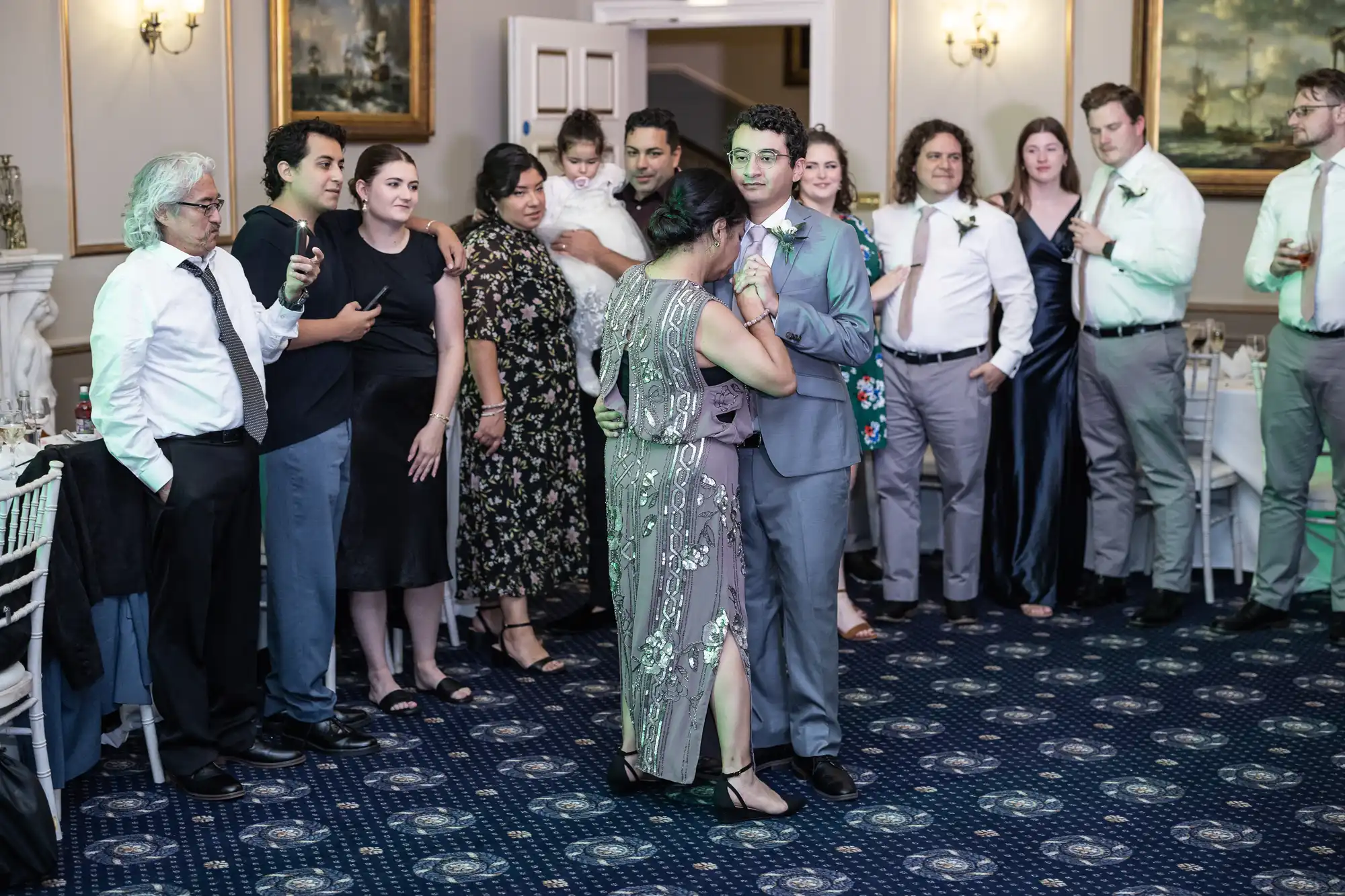 A group of people, dressed formally, stand on a dark blue patterned carpet in a well-lit room. Two individuals in the center, one in a suit and the other in an embellished dress, engage in a close embrace.