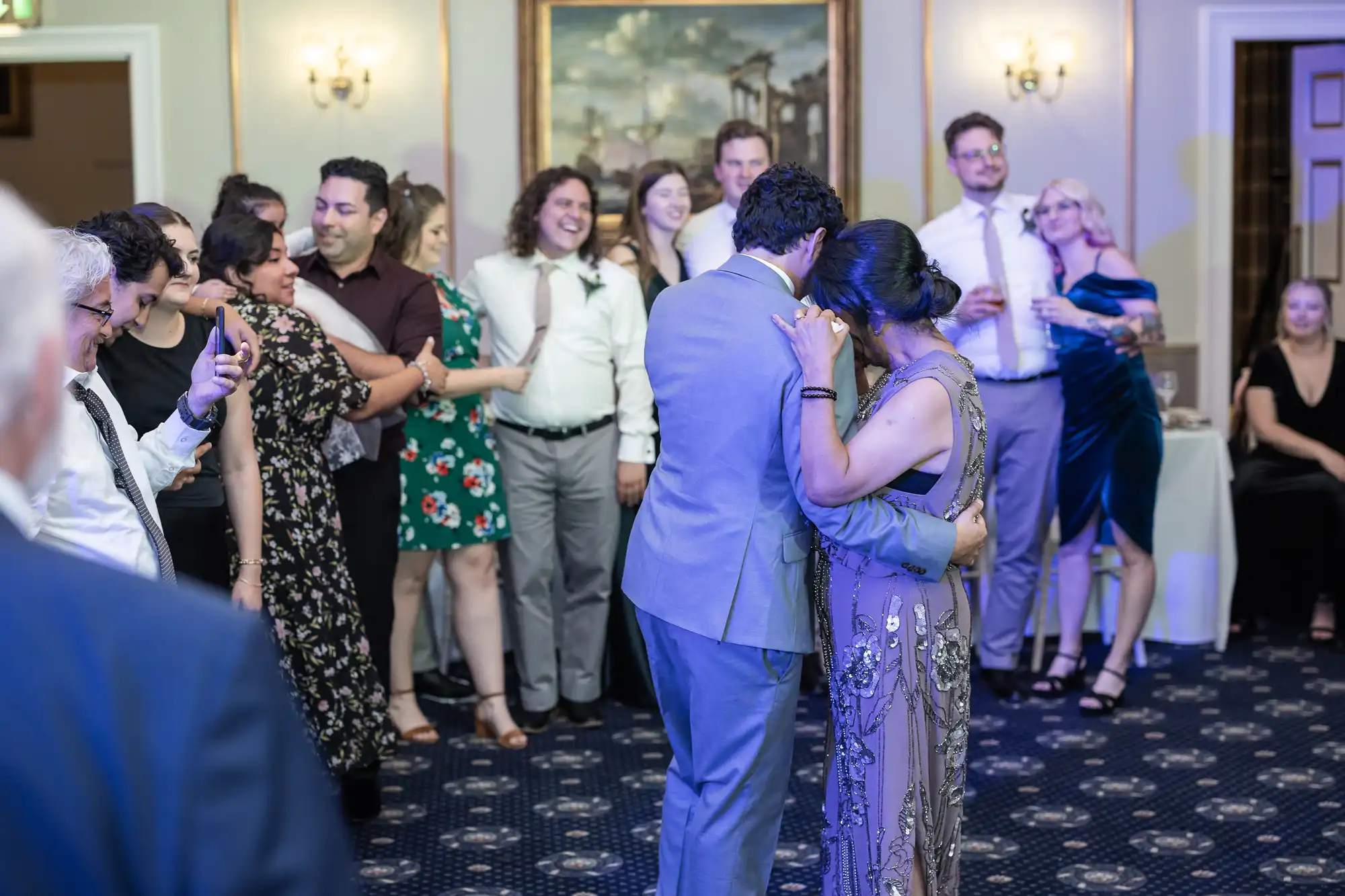 Couple in formal attire dancing, surrounded by a group of people watching and smiling in a room with ornate decor.