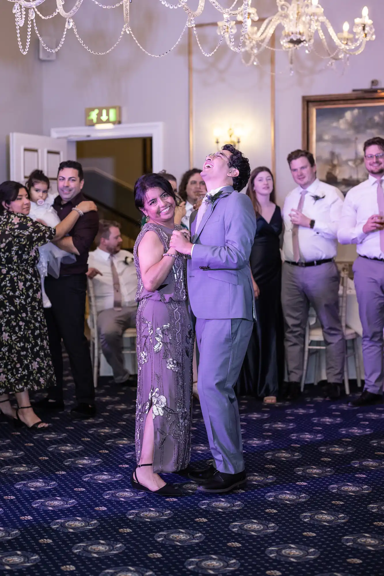 A woman in a floral dress and a man in a grey suit dance together in a decorated room, surrounded by smiling onlookers and a chandelier overhead.