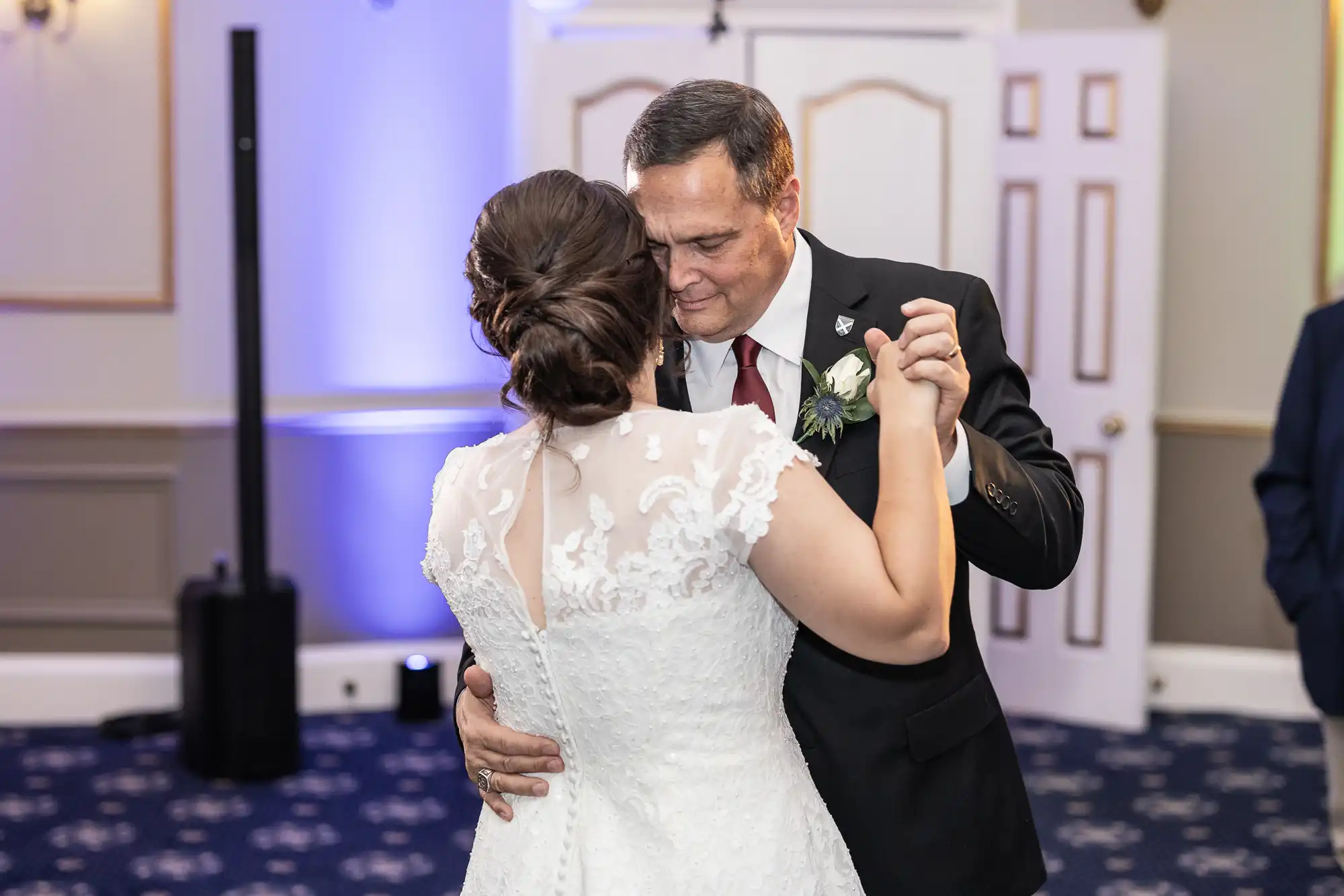 A bride in a white wedding dress and a man in a black suit are dancing together in an indoor venue with a blue and white patterned carpet. Their heads are close as they share a moment.