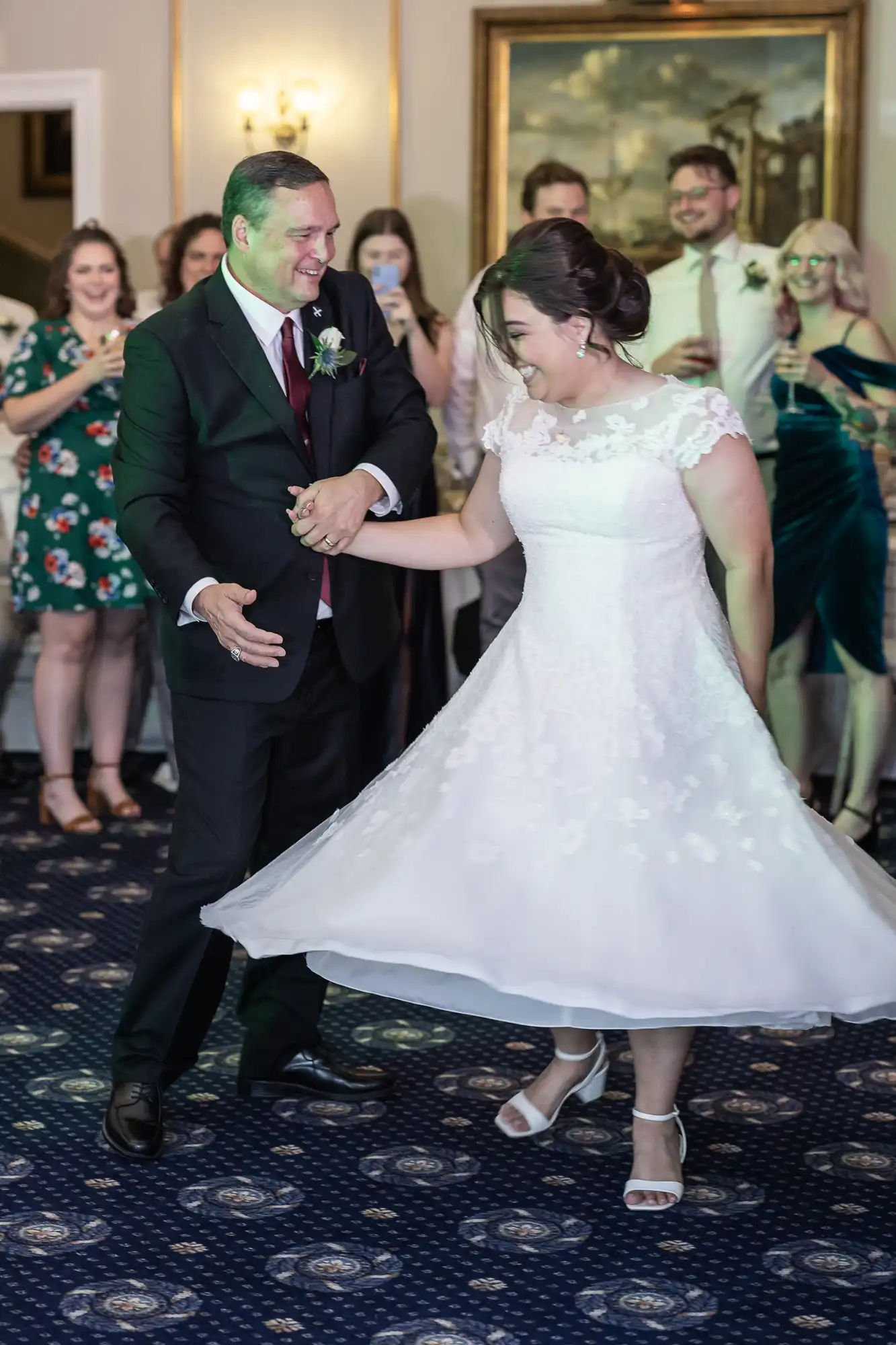 A man in a suit and a woman in a white dress dance together at a wedding celebration, with guests watching and smiling in the background.