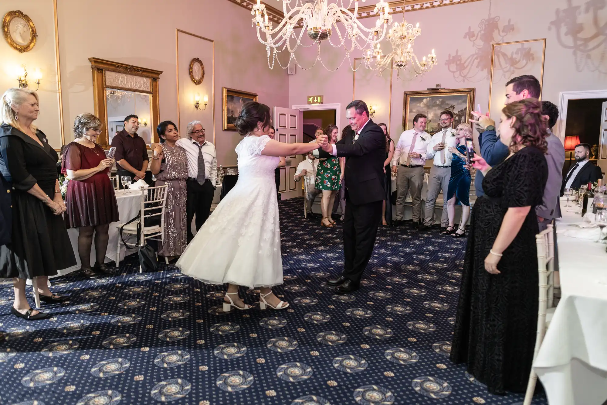 A bride and groom dance in the center of a room surrounded by standing guests in formal attire, under chandeliers.