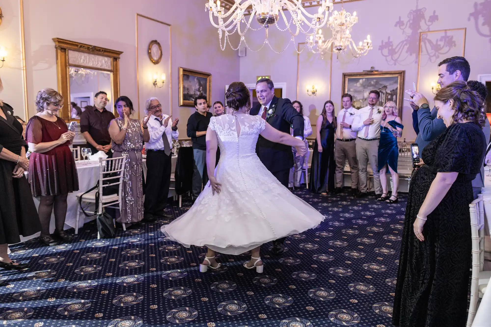 A couple dances in the center of a room at their wedding reception while guests stand around clapping and taking photos. The bride wears a white dress, and the groom wears a suit.