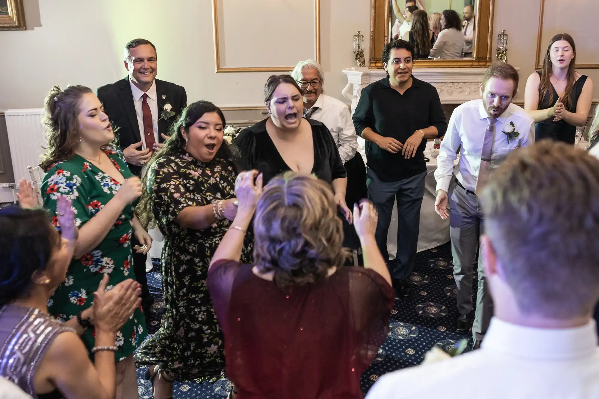 A group of people dressed in formal attire are dancing and clapping hands at a social event indoors. Some are smiling and laughing, while others look engaged in conversation.