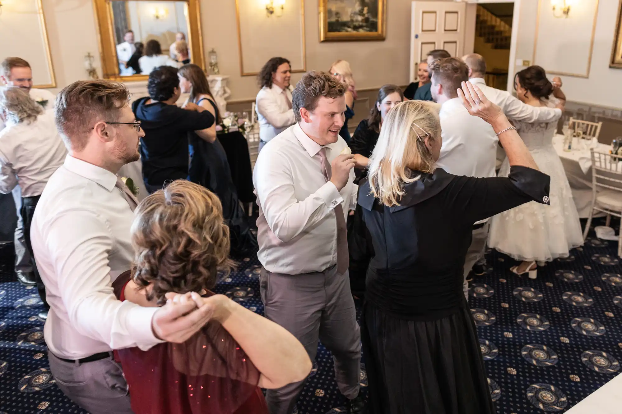 Group of people dancing and mingling at an indoor event, with some dressed in formal attire. The space is decorated with mirrors, lights, and framed artwork on the walls.