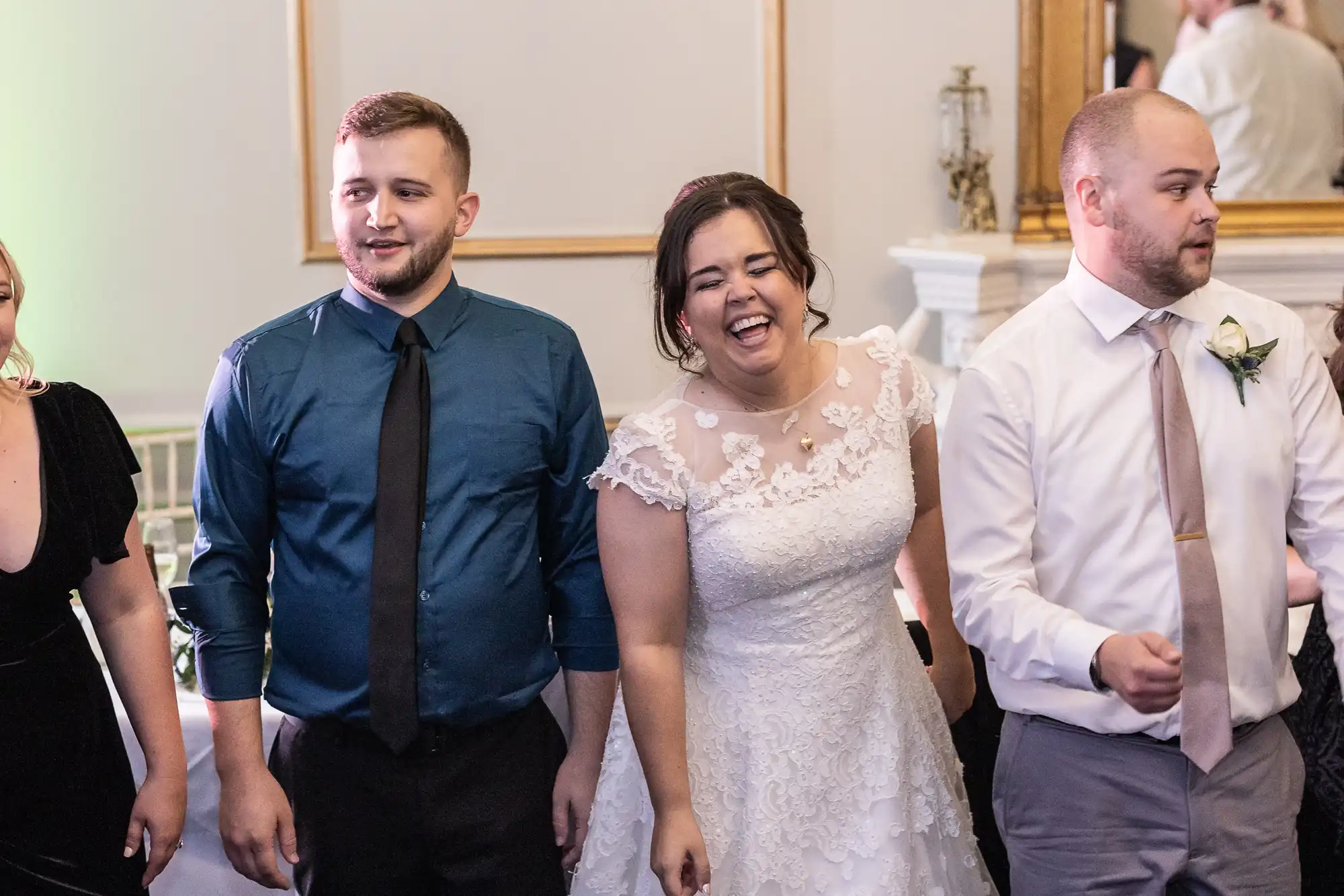 A bride in a white lace dress stands between two men with neckties, one in a blue shirt and the other in a white shirt. They are indoors, and she is smiling widely.