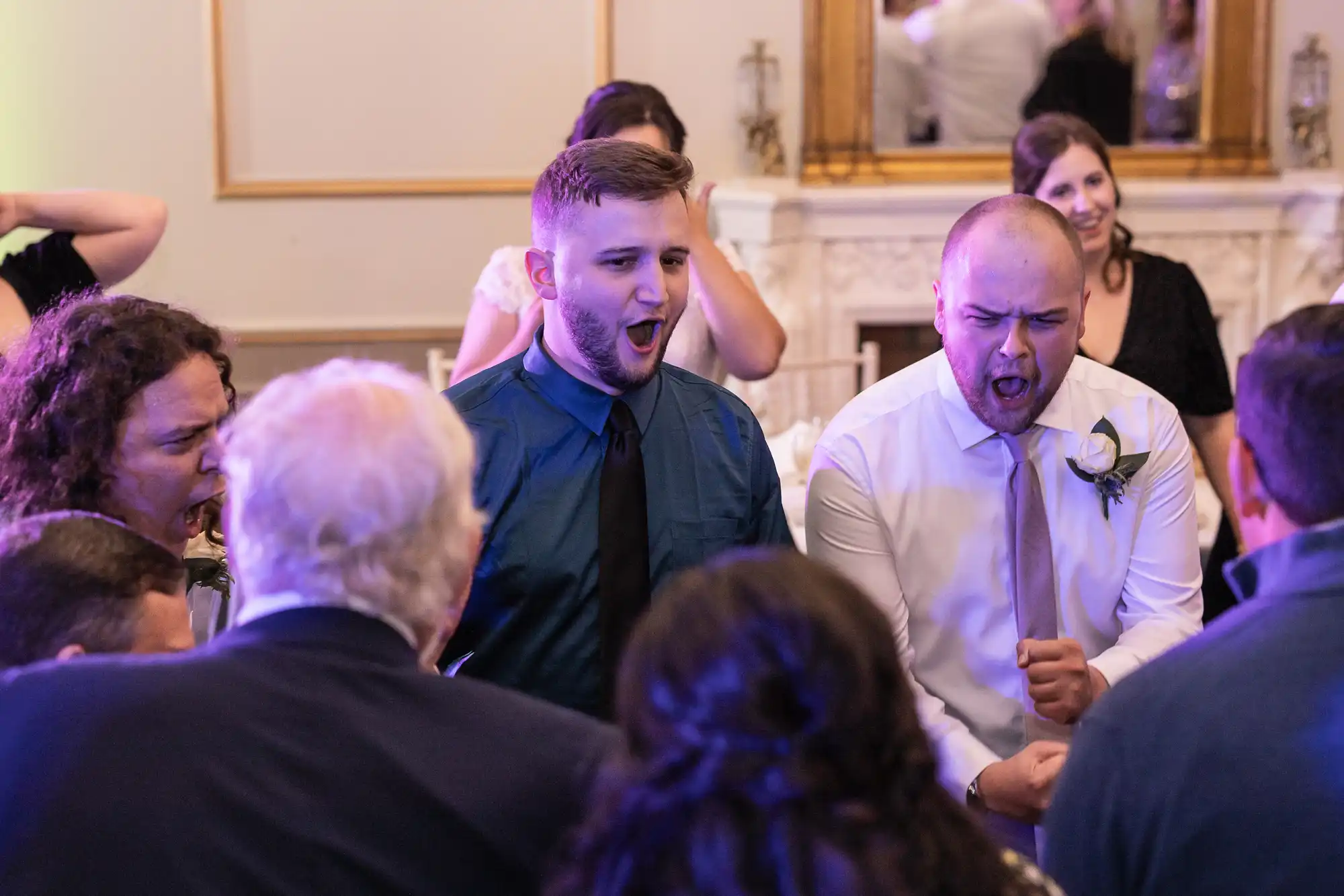 Group of people in formal attire enthusiastically singing or shouting together in a festive setting with a mirror and fireplace in the background.