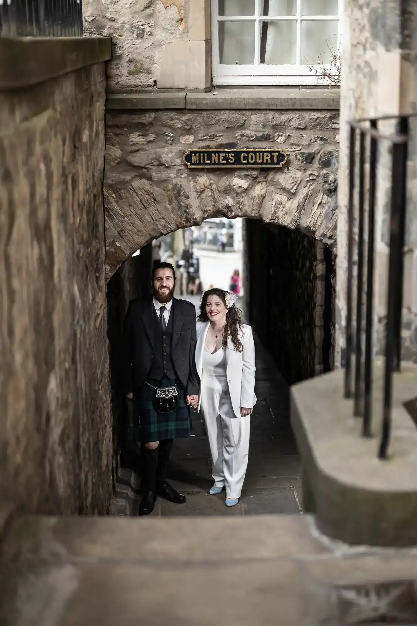 A man in a kilt and a woman in a white suit stand holding hands under a stone archway labeled "Milne's Court" in an old alleyway.