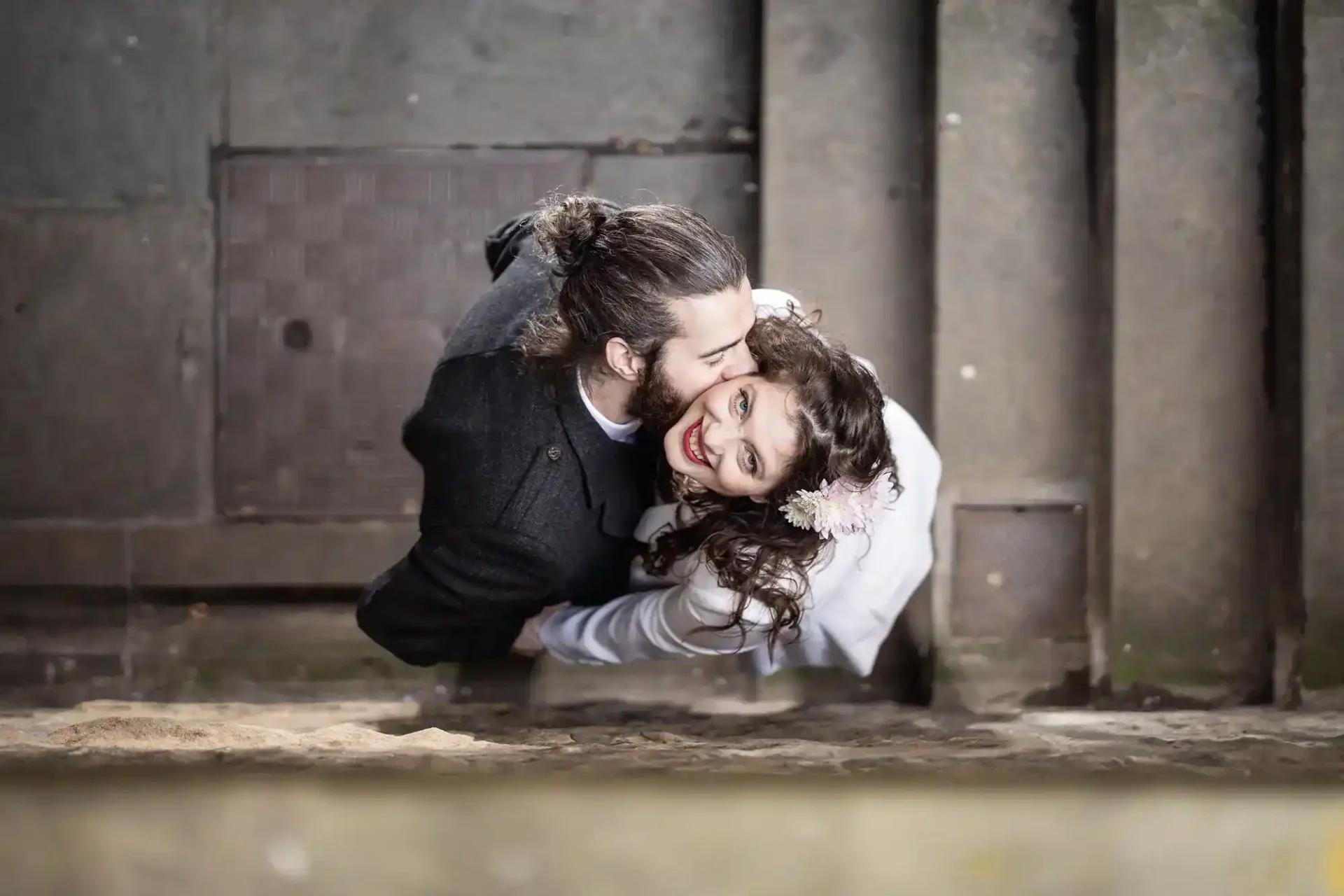 An overhead view of a man kissing a smiling woman on the cheek as they embrace, standing on a stone surface.