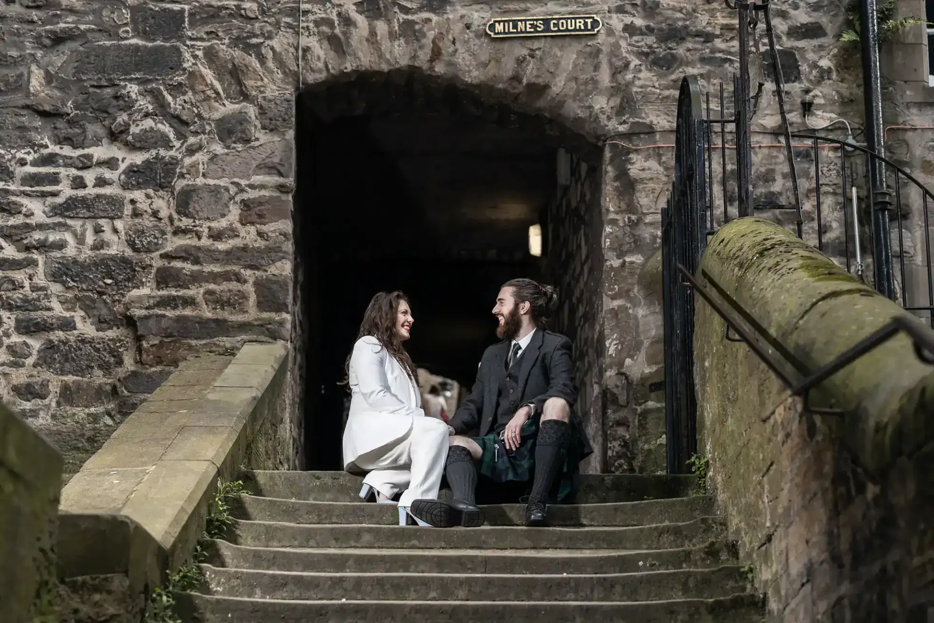 A woman in a white coat and a man in traditional Scottish attire sit together on stone steps under an archway labeled "Milne's Court.