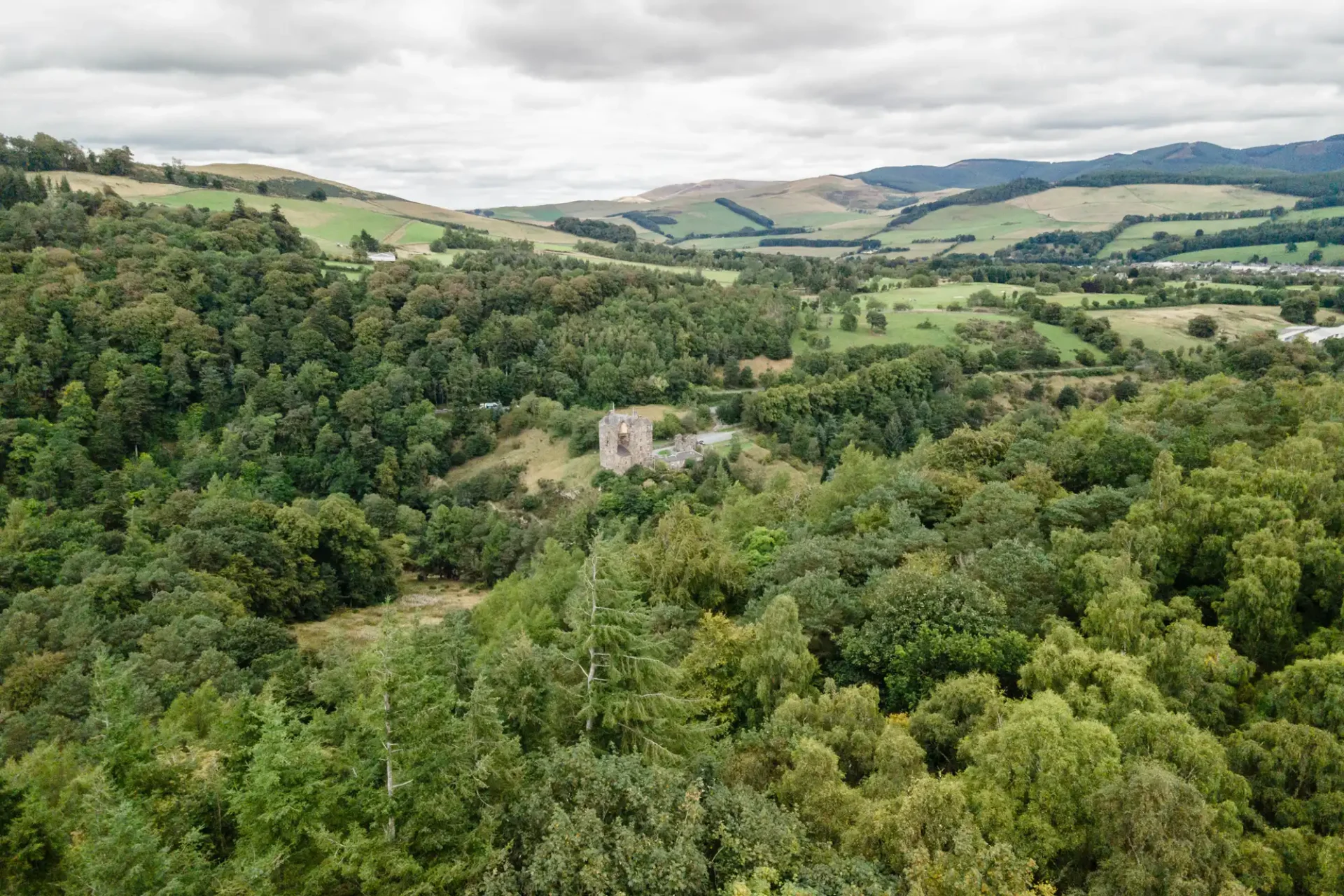 Image by Neidpath Castle photographer of the aerial view of an ancient stone tower surrounded by dense forest, with rolling hills and scattered farmland in the background under a cloudy sky.