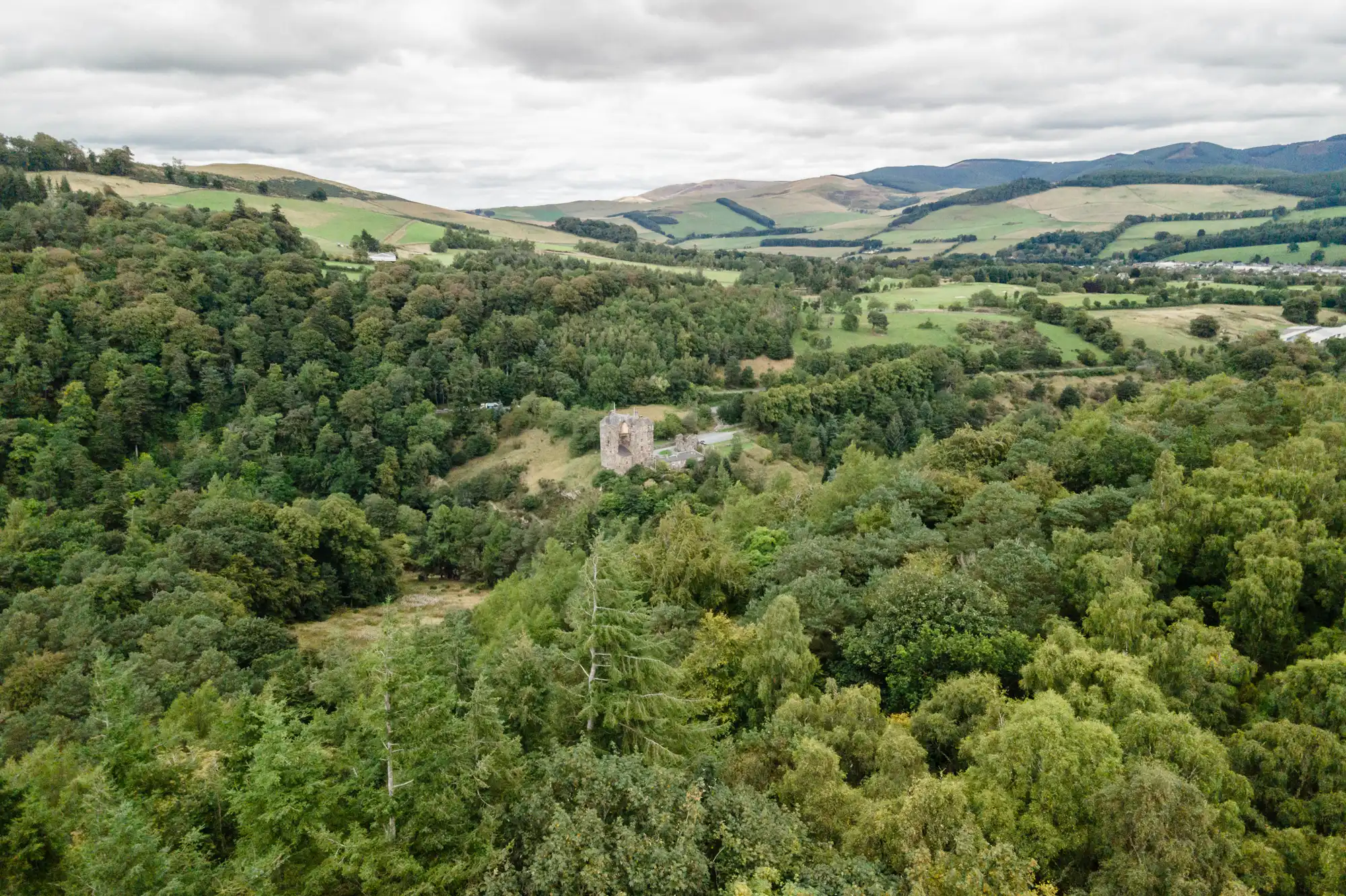 Aerial view of an ancient stone tower surrounded by dense forest, with rolling hills and scattered farmland in the background under a cloudy sky.