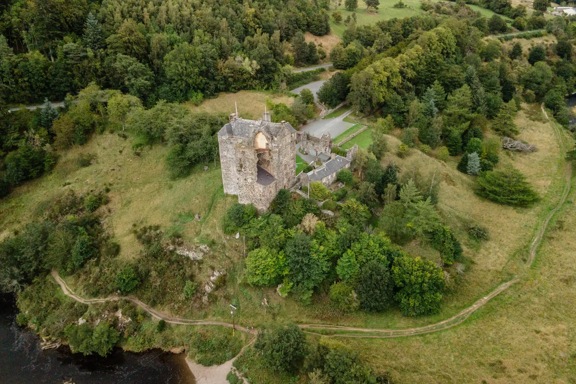 Aerial view of a partially ruined stone castle surrounded by greenery and trees, situated on a hill above a river, with walking paths nearby.