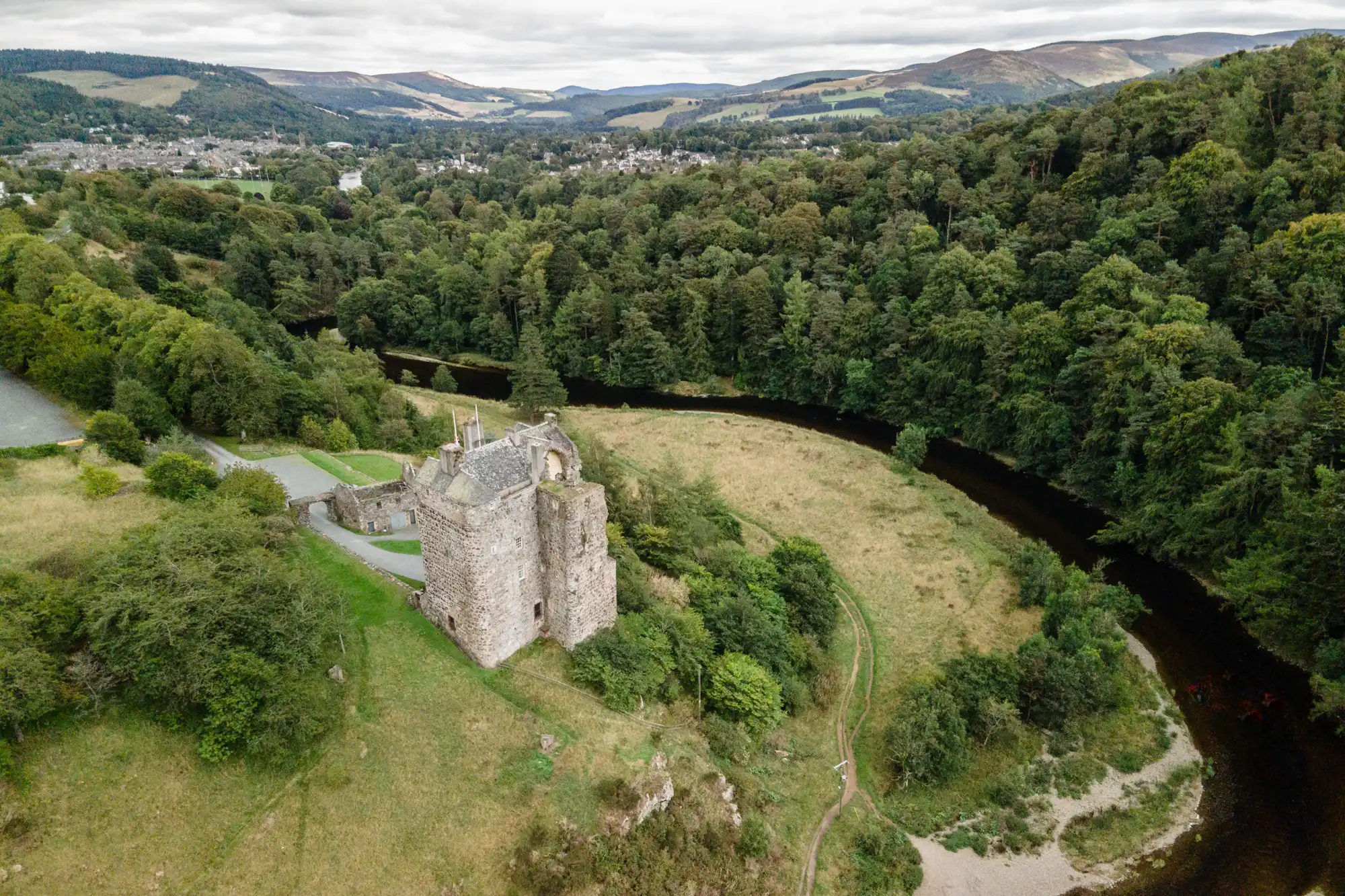 Aerial view of a partially ruined stone castle surrounded by lush greenery, with a river flowing nearby and hills in the background.