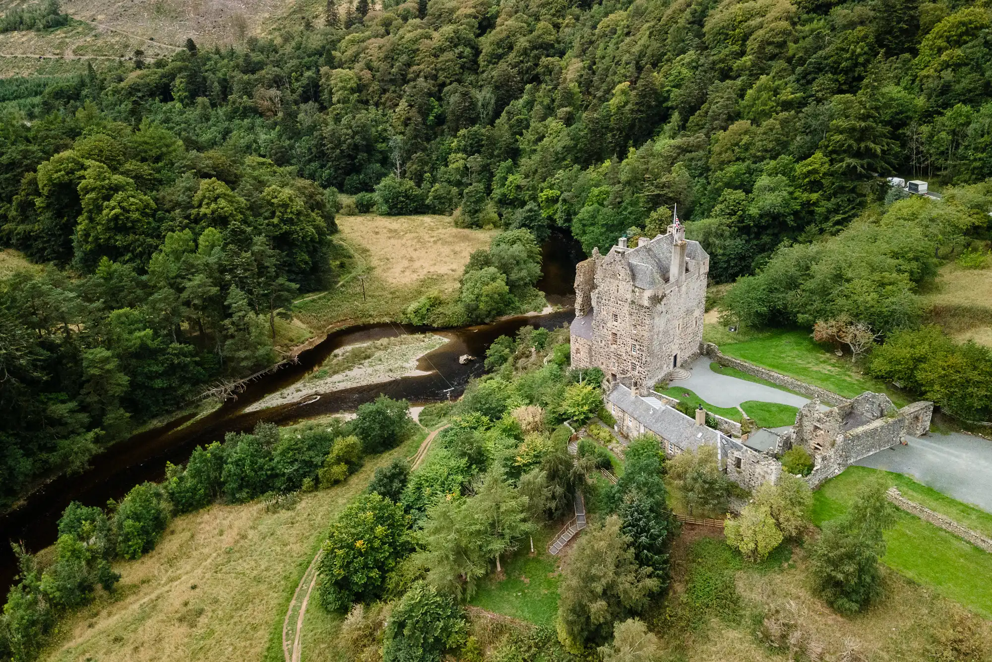 Aerial view of a historical stone castle surrounded by lush greenery and a winding river in a forested landscape.