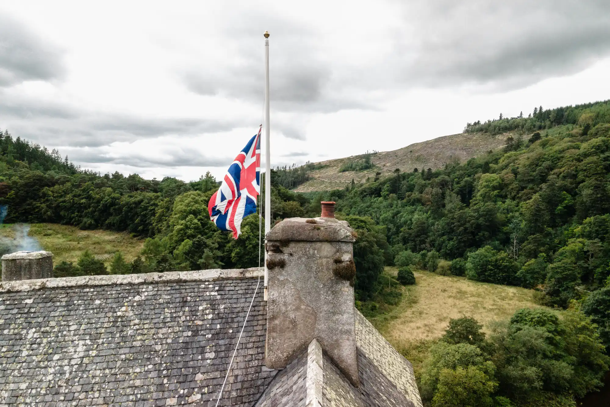 A Union Jack flag is at half-mast on a stone building's rooftop, set against a backdrop of rolling green hills and cloudy sky.