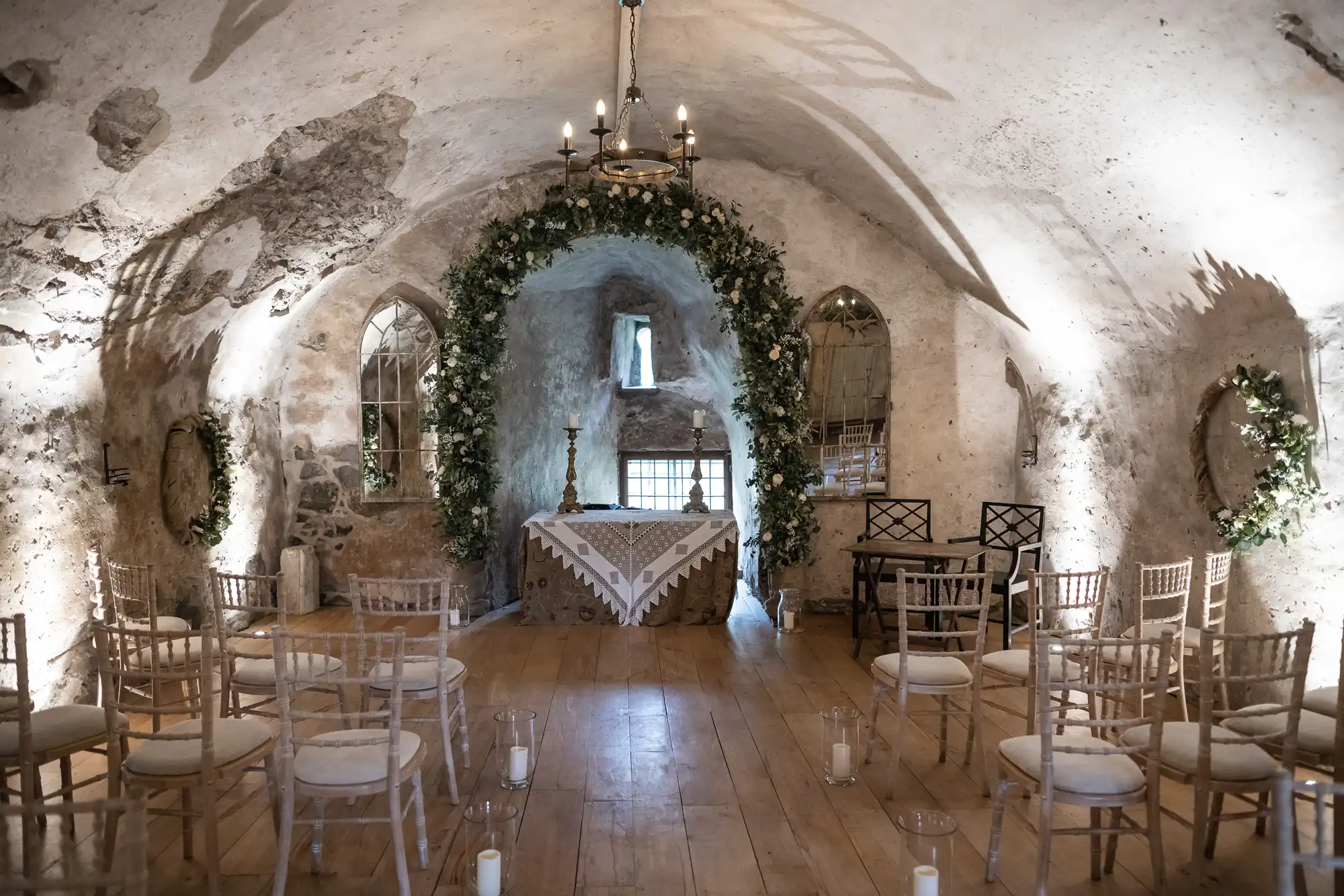 A small, rustic chapel with stone walls and a wooden floor is decorated with floral arches and white chairs arranged for a ceremony. There's an altar at the front under soft lighting.