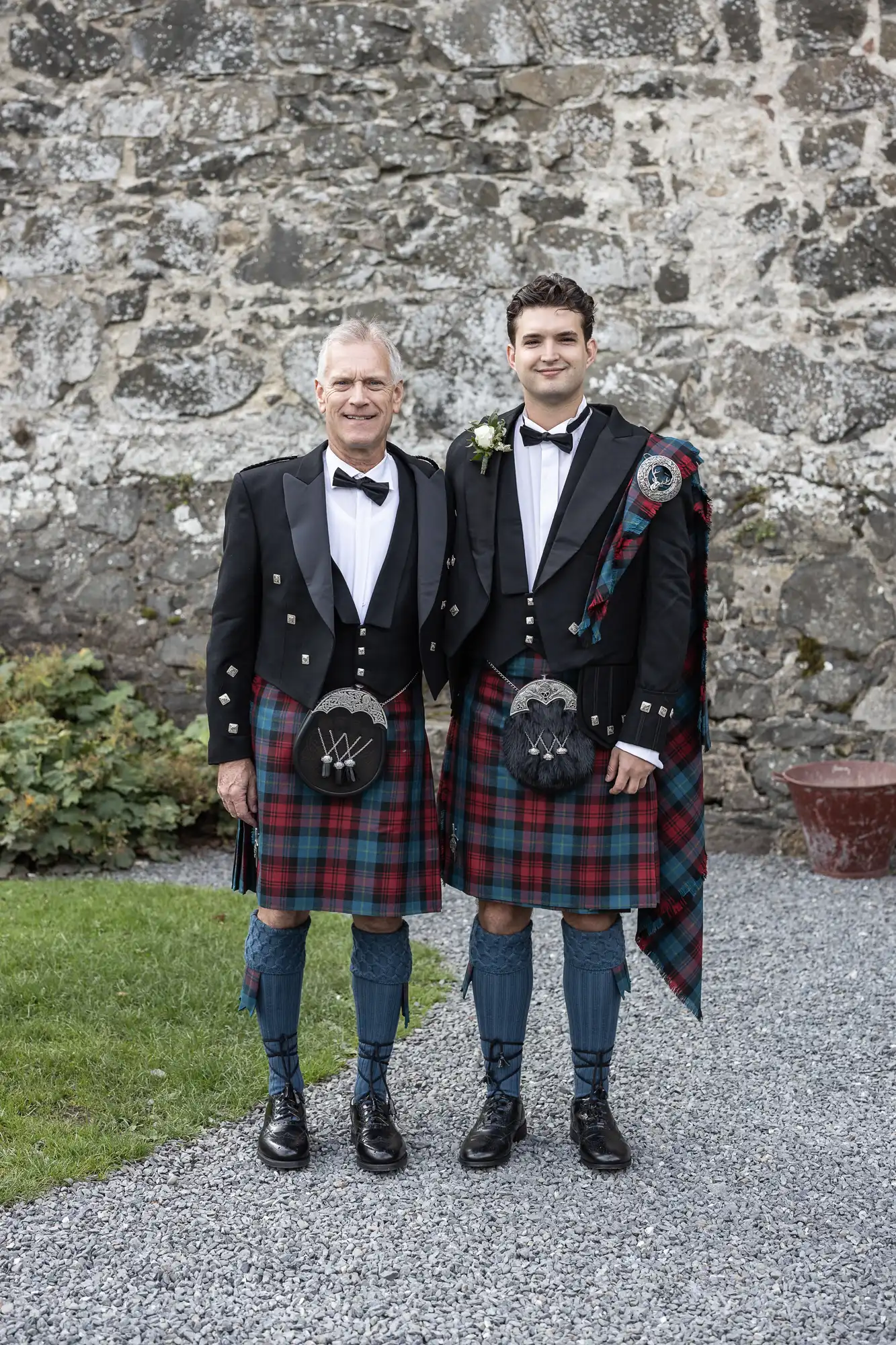 Two men dressed in traditional Scottish kilts stand side by side outdoors in front of a stone wall, smiling at the camera.