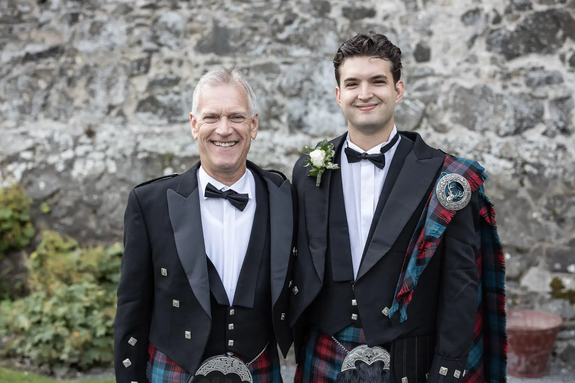 Two men in formal Scottish attire with black jackets, bow ties, and tartan kilts pose in front of a stone wall. The younger man wears a sash with a floral pin on the lapel.