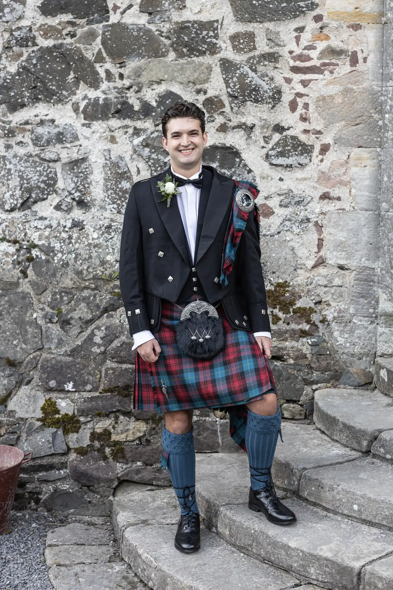 A man stands on stone steps in front of a stone wall, wearing traditional Scottish attire including a kilt, sporran, and a jacket with a boutonnière.