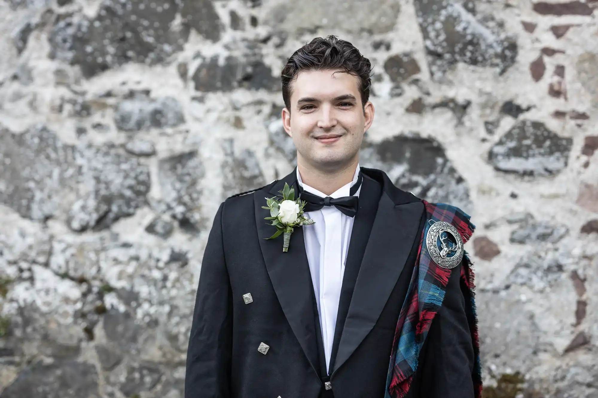 A man in formal attire stands in front of a stone wall, wearing a black jacket with buttons, a white shirt, a bow tie, and a tartan sash. He is also adorned with a white boutonniere.