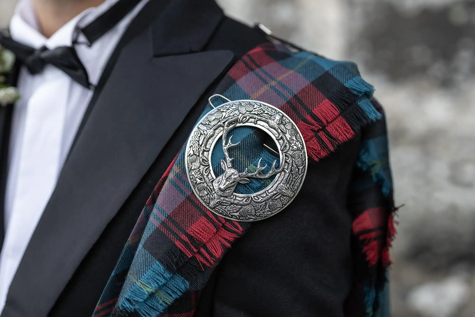 Close-up of a person in formal attire wearing a black jacket with a tartan sash and a silver brooch featuring a stag's head.