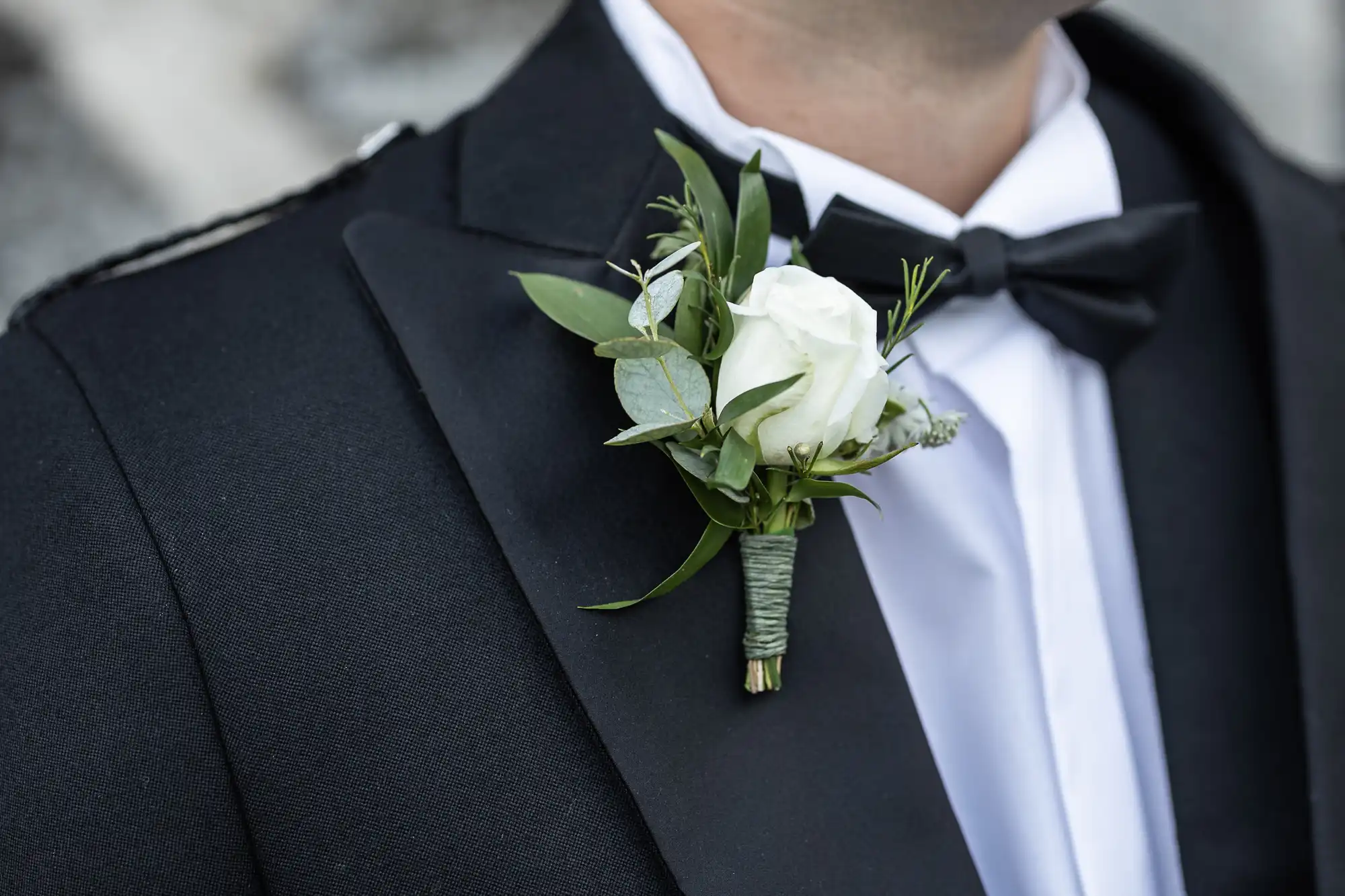 Close-up of a person wearing a black suit jacket, white shirt, and black bow tie, with a boutonnière made of a white rose and greenery pinned to their lapel.