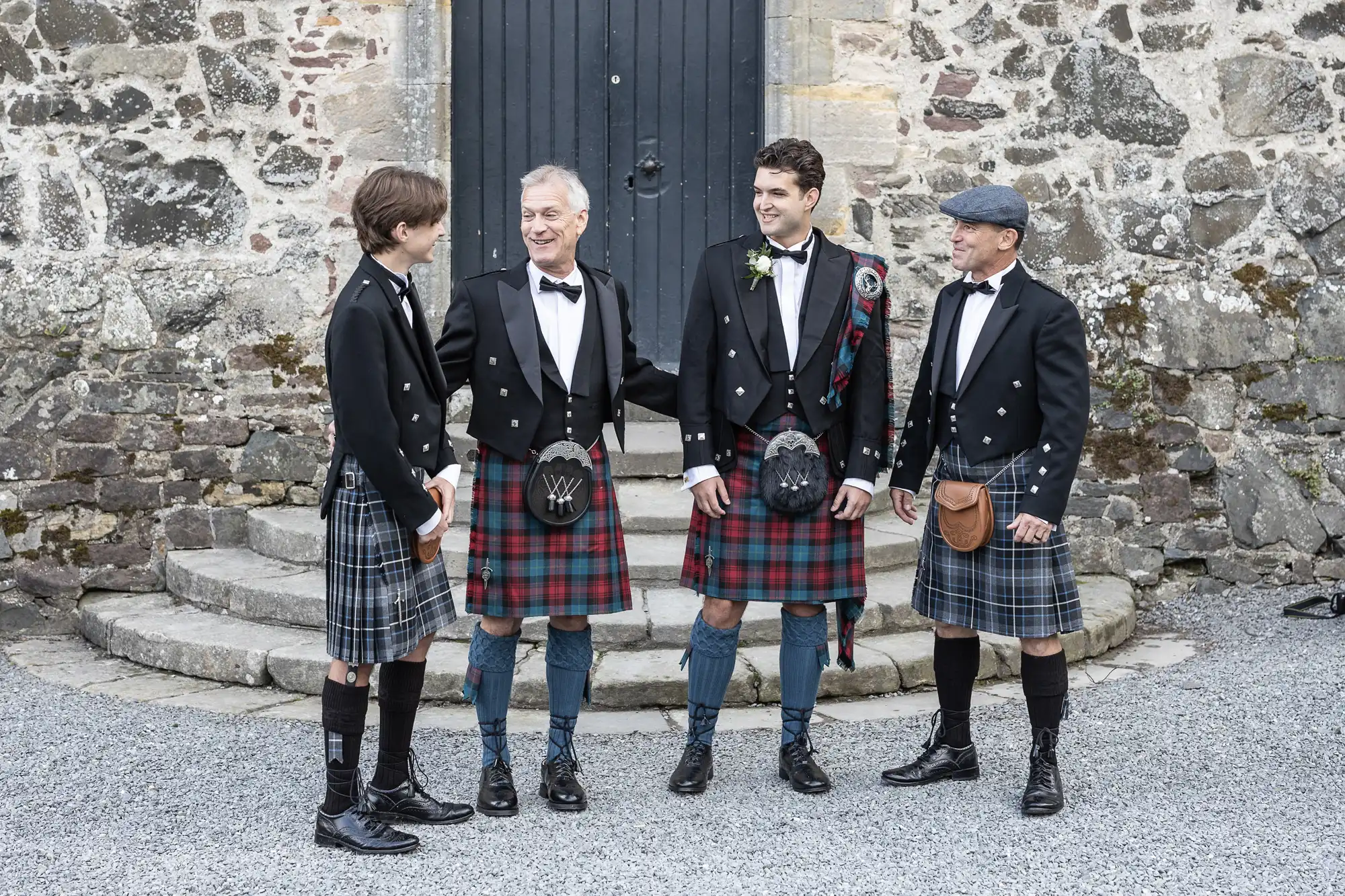 Four men in kilts stand in front of a stone building, conversing and smiling.