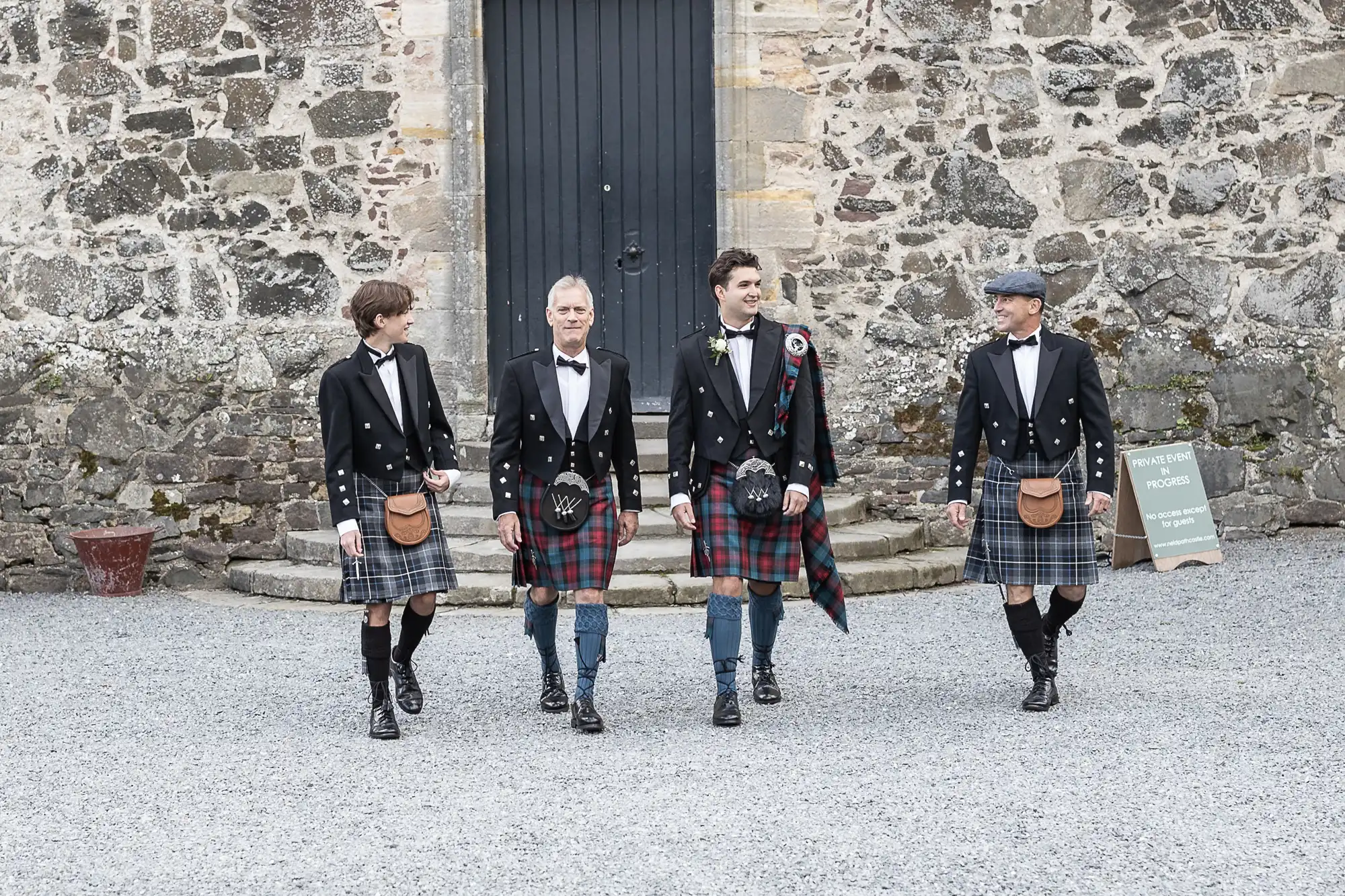 Four men in kilts and formal jackets walk together in front of a stone building.