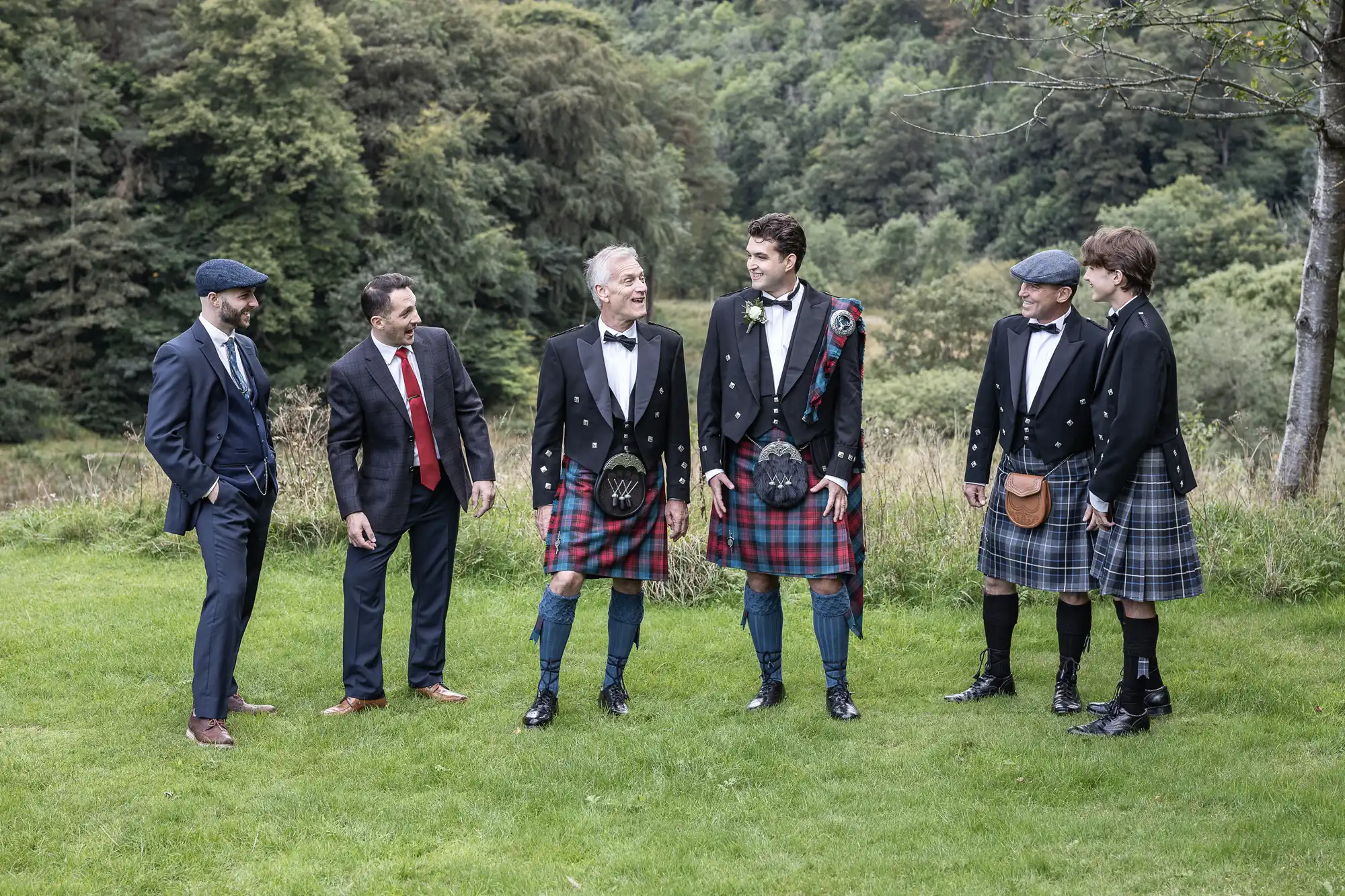 Five men standing on grass, four in kilts, one in a suit. Plaid kilts, jackets and ties. Background features trees and greenery. They appear engaged in conversation, with a jovial atmosphere.