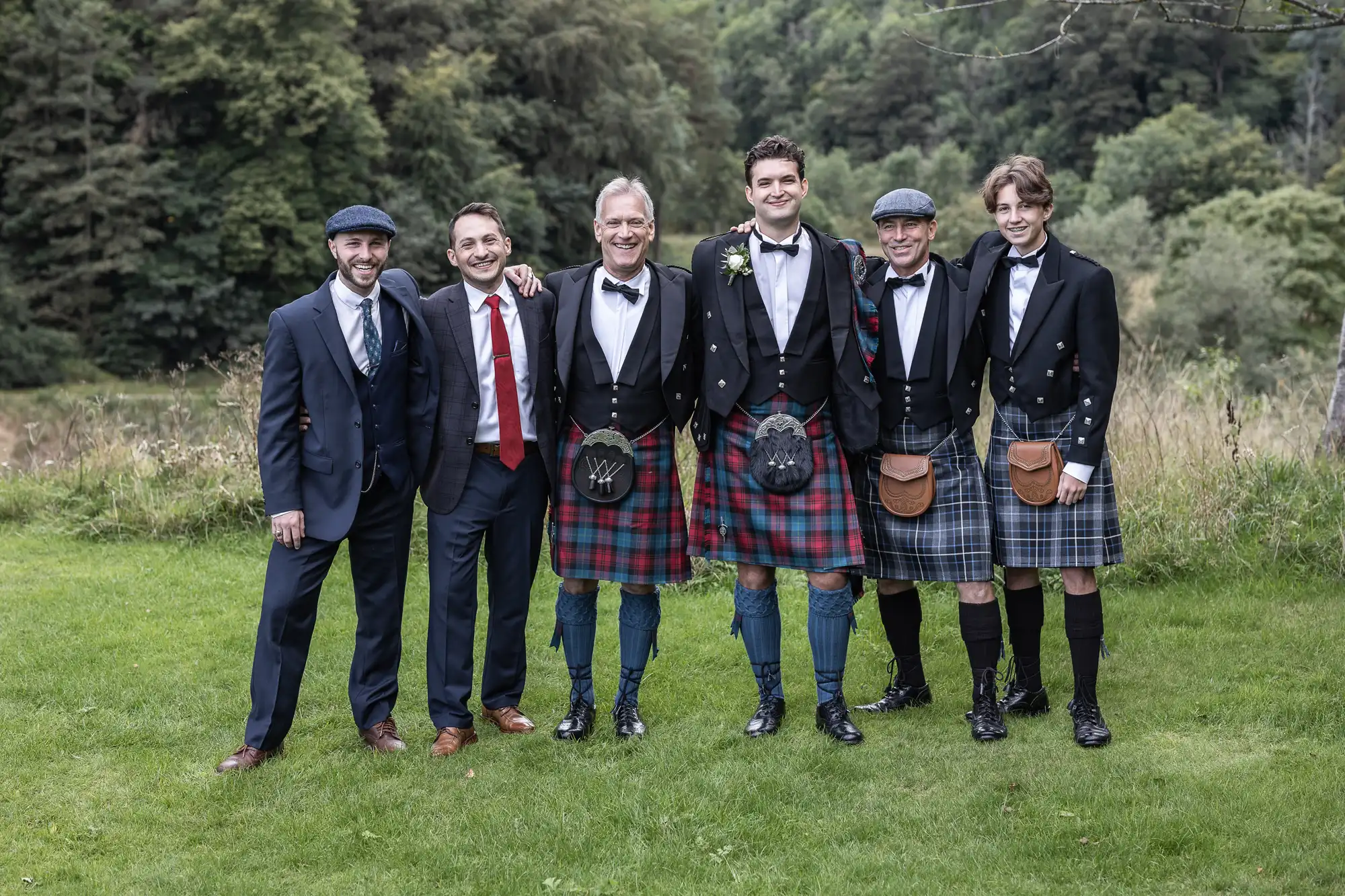 Six men in formal attire pose outdoors, four wearing kilts and two in suits, with trees and grass in the background.