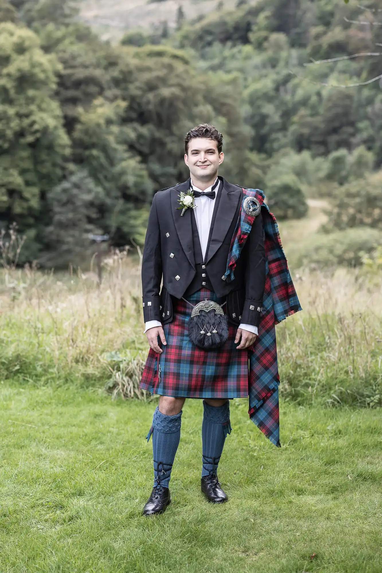 A person wearing a traditional Scottish kilt outfit stands on grass, with trees and hills in the background.