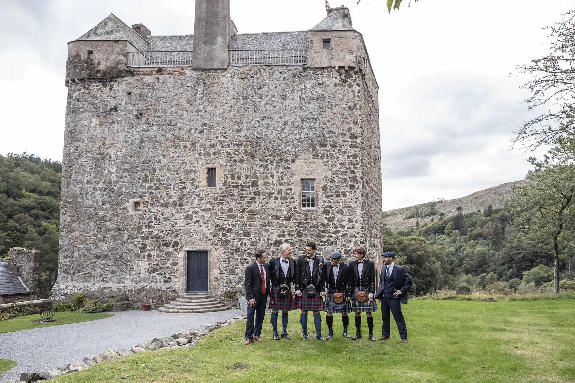 A group of men standing in kilts and suits in front of a stone castle with a large chimney, surrounded by grass and scattered trees.