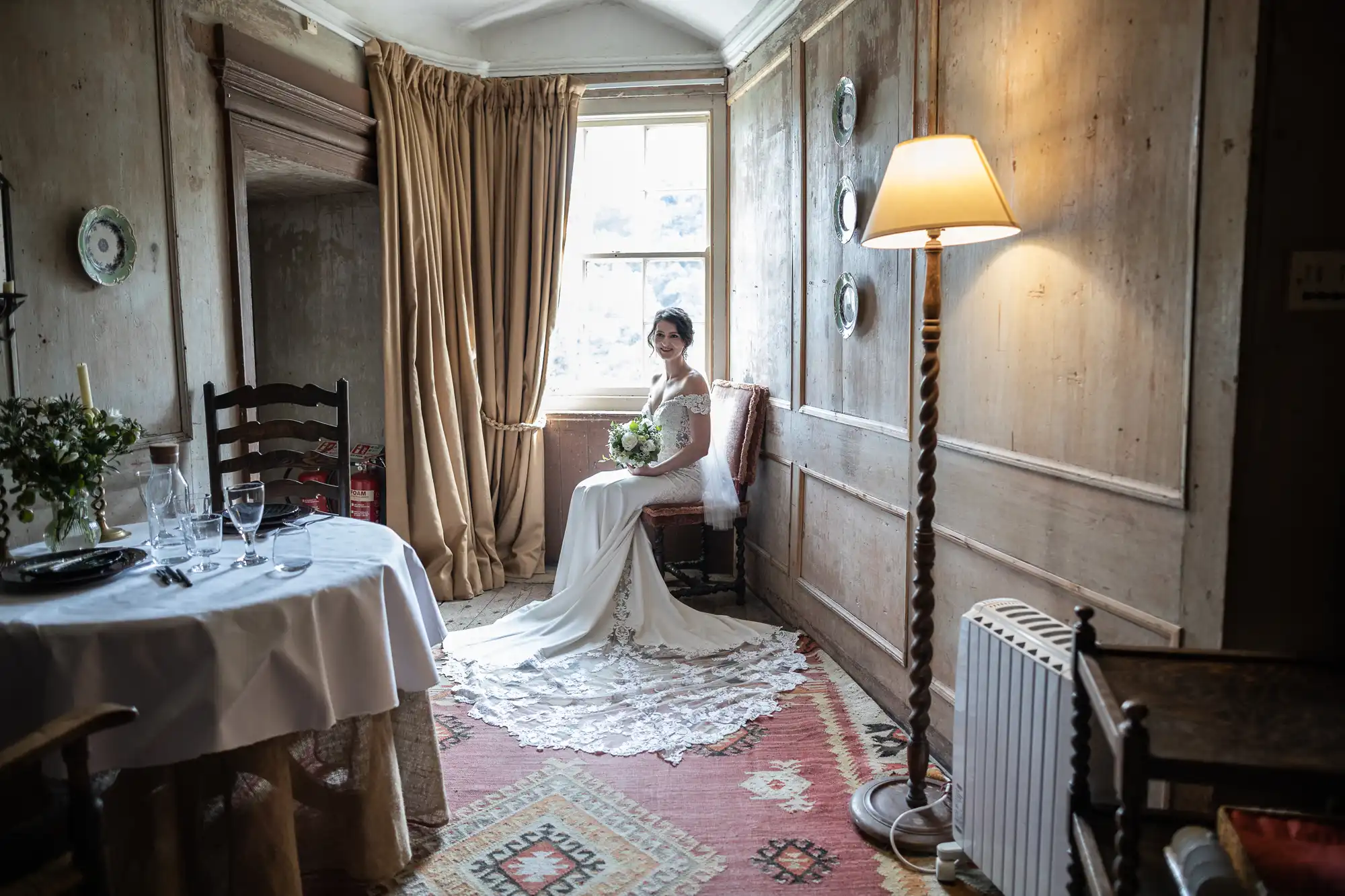 A bride in a white dress sits by a window holding a bouquet in a rustic room with wooden walls, a dining table set for two, and a lit floor lamp nearby.