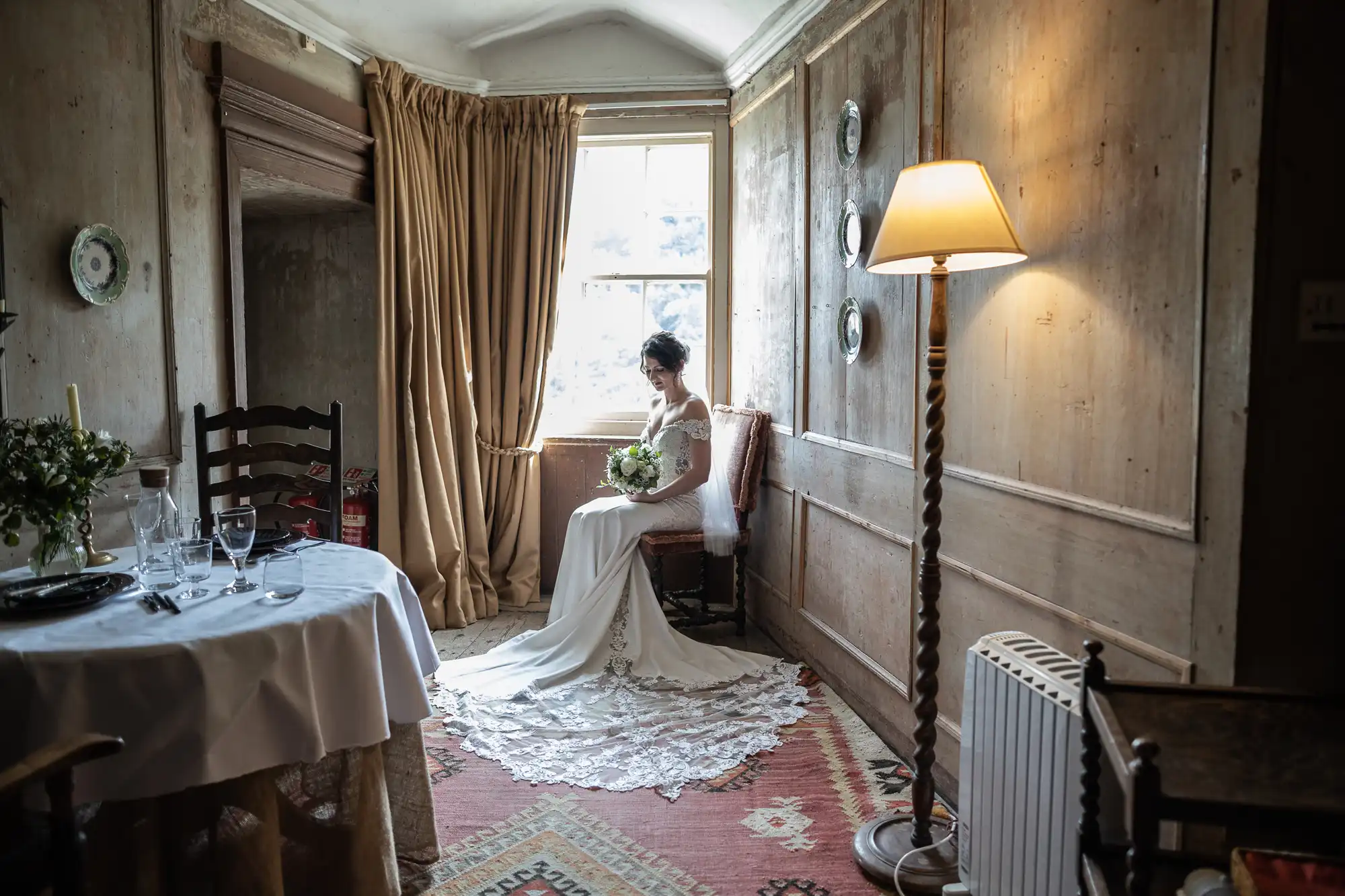 Bride in a white gown sitting near a window holding a bouquet in a rustic room with wooden walls, a table set for dining, a floor lamp, and a radiator on a patterned rug.