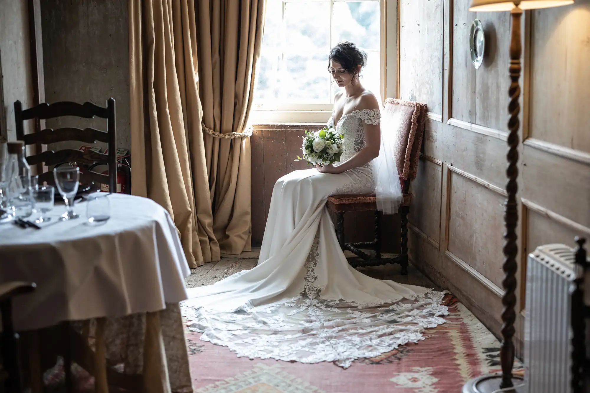 A bride in a white gown sits on a chair by a window holding a bouquet, with a long lace train spread on the floor. The room has wooden paneling, a table with glassware, and a curtain drawn open.