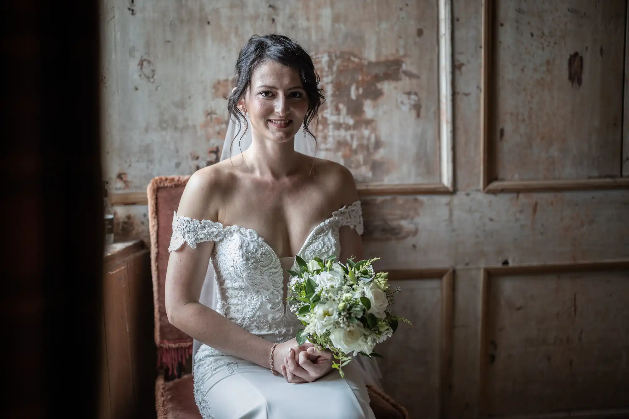 A bride in a white lace wedding dress holds a bouquet of white and green flowers while sitting on a chair, with a rustic wooden wall in the background.