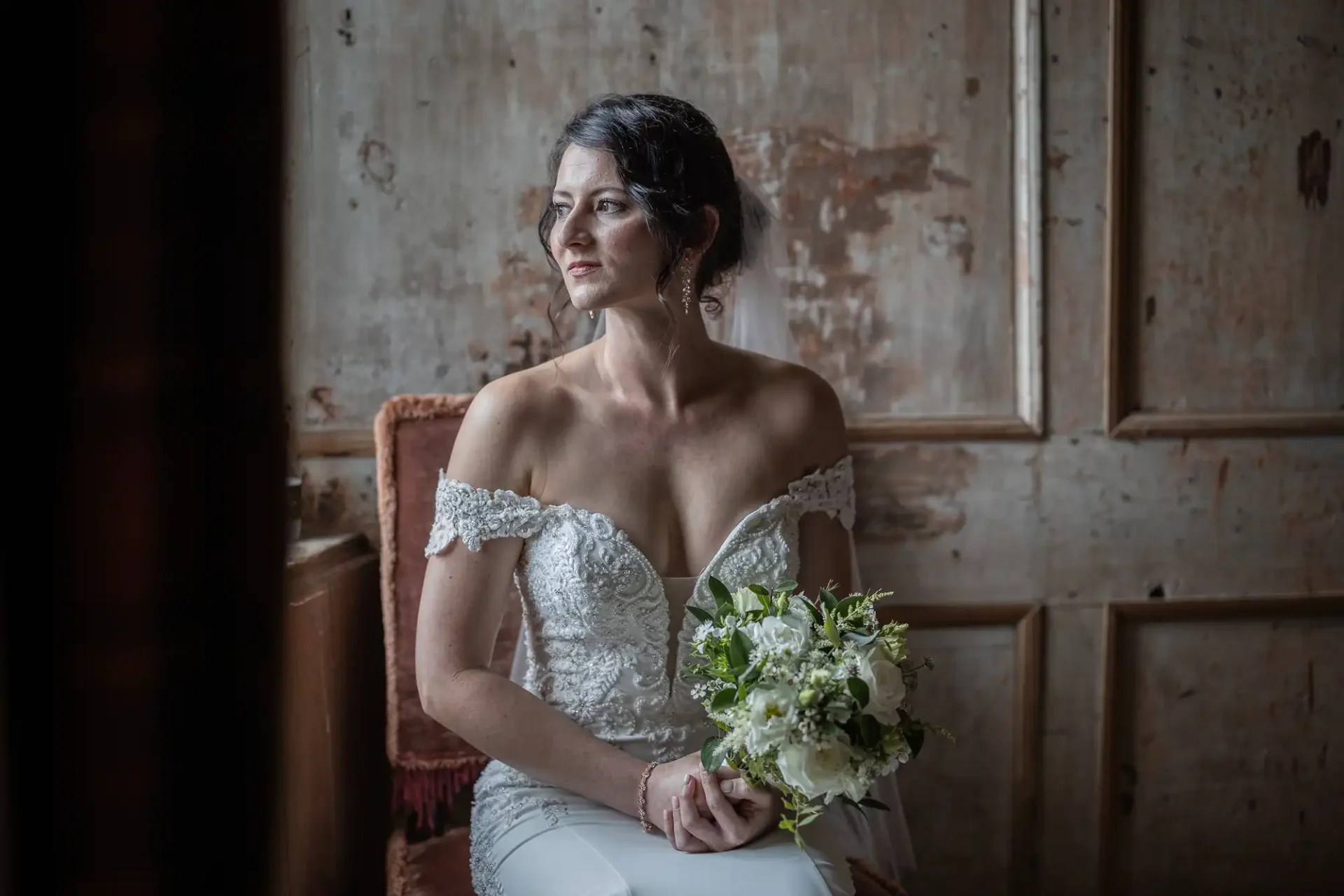 A bride in a white, off-shoulder wedding dress holds a bouquet while sitting on a chair, looking to the side against a rustic, textured background.