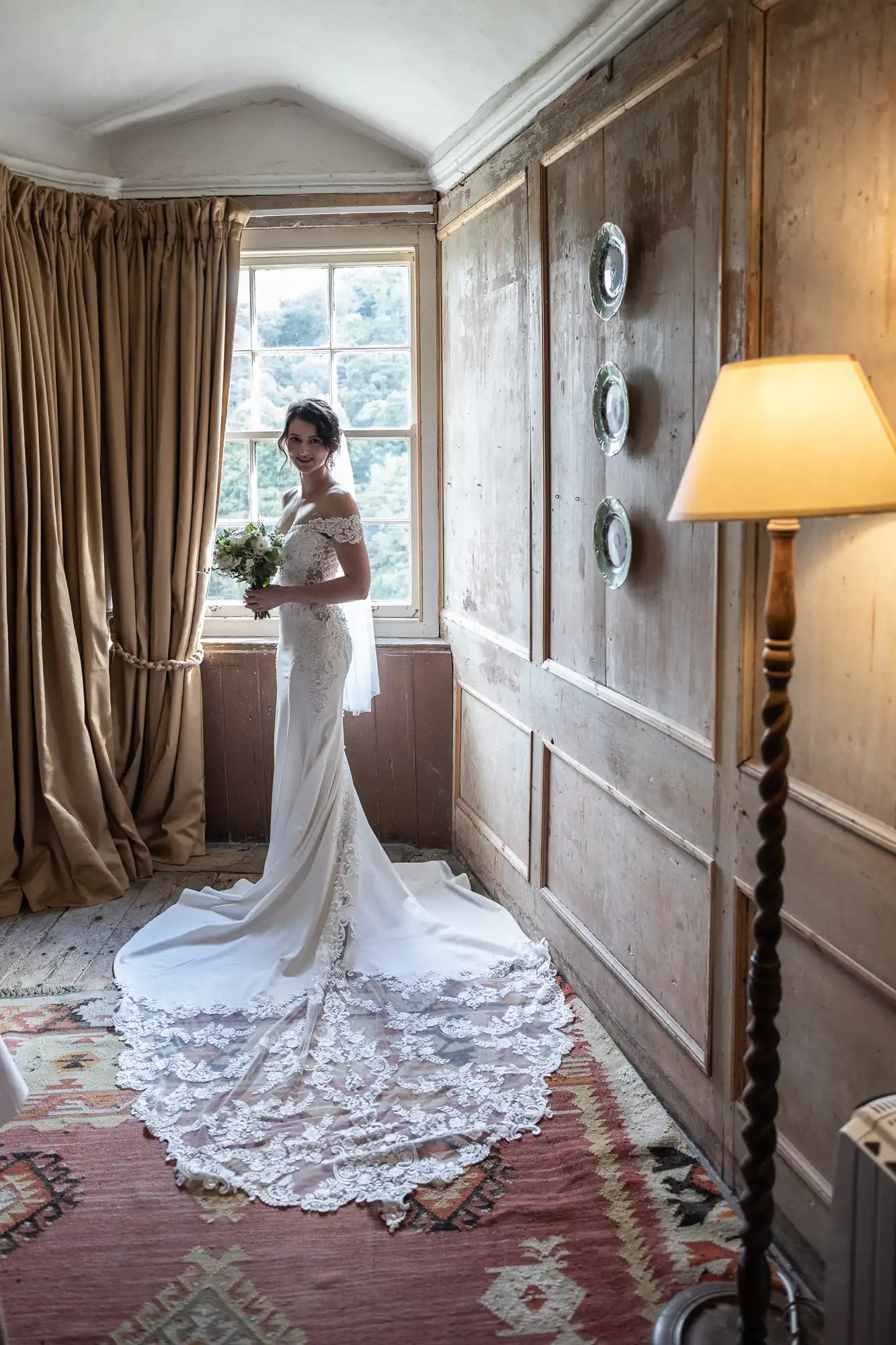 A bride in a white wedding dress with a long lace train stands near a window in a wooden-paneled room, holding a bouquet and smiling.