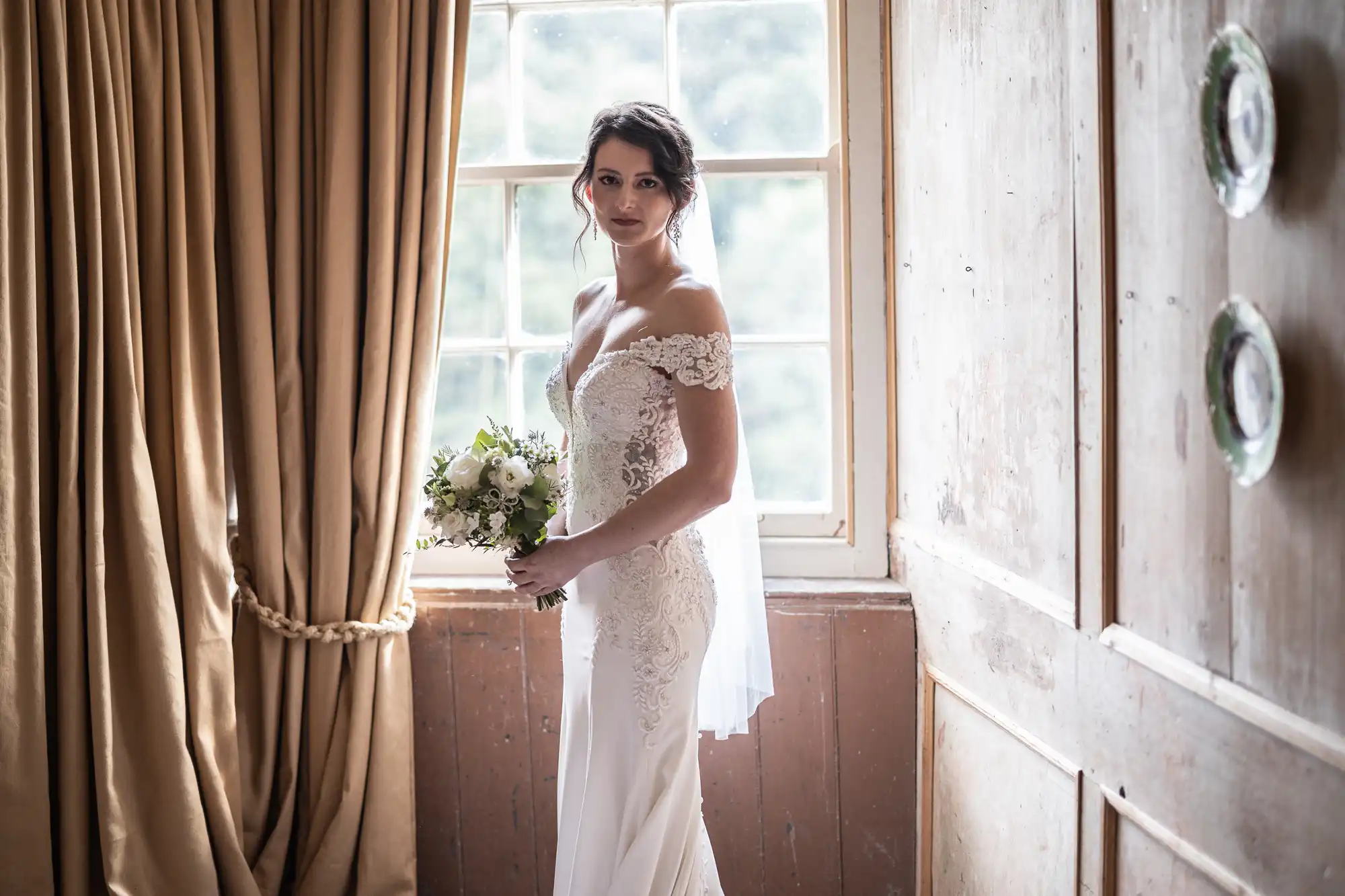 A bride in a white lace off-shoulder gown stands by a window holding a bouquet of flowers. Tan curtains are pulled back, and decorative plates are mounted on a wooden wall.