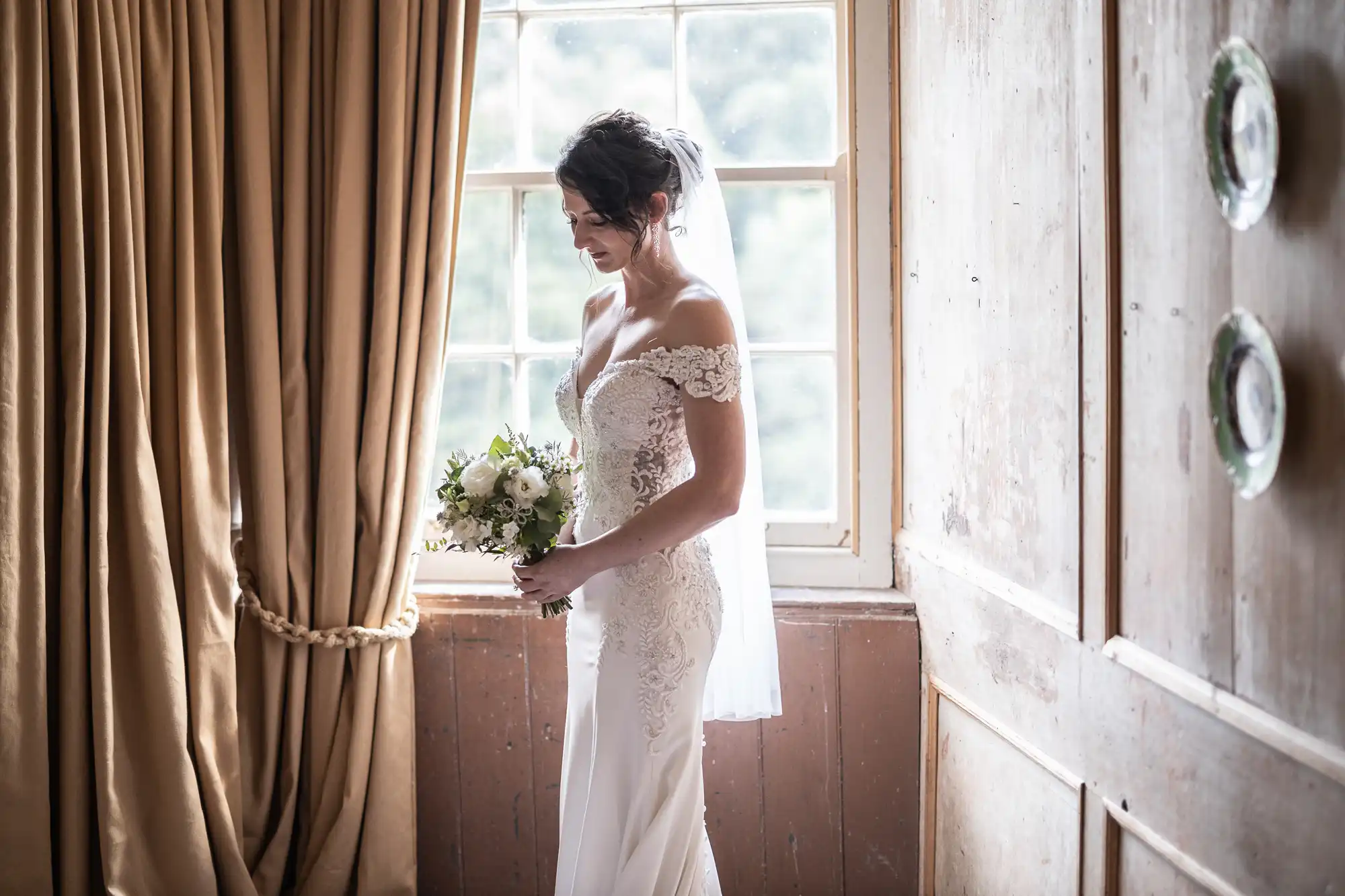 A bride in a lace wedding gown holds a bouquet while standing by a window with golden curtains. Light pours in, illuminating her figure.