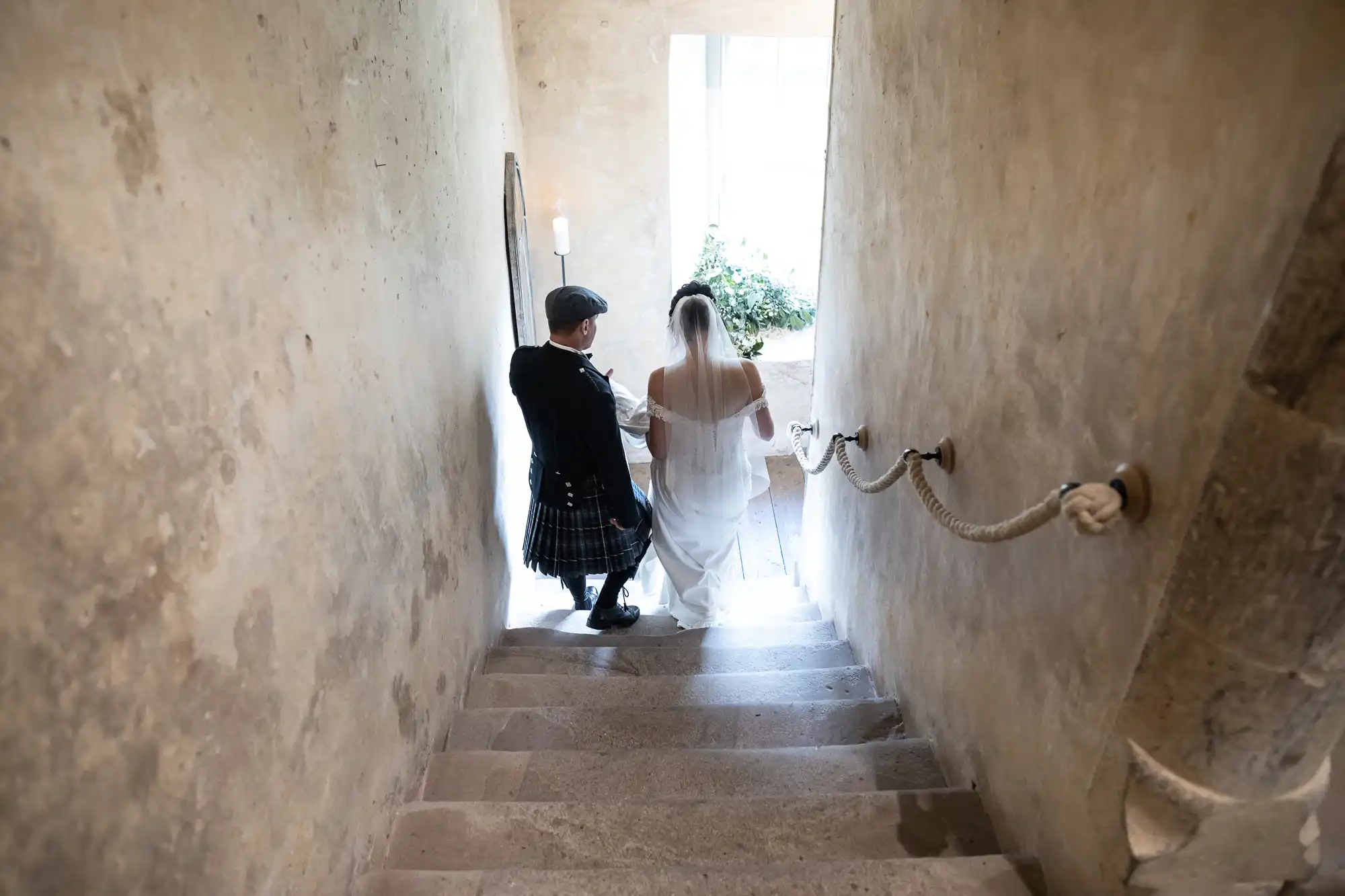 A couple dressed in wedding attire, with the groom in a kilt and the bride in a white gown, walk down a set of stone stairs in a narrow, arched hallway.