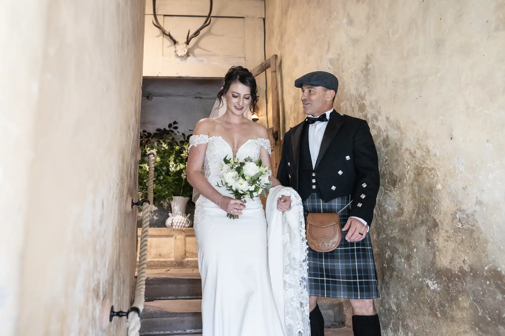 Bride in a white dress with a bouquet descends stone stairs, accompanied by a man in traditional Scottish attire, with a beige wall backdrop.