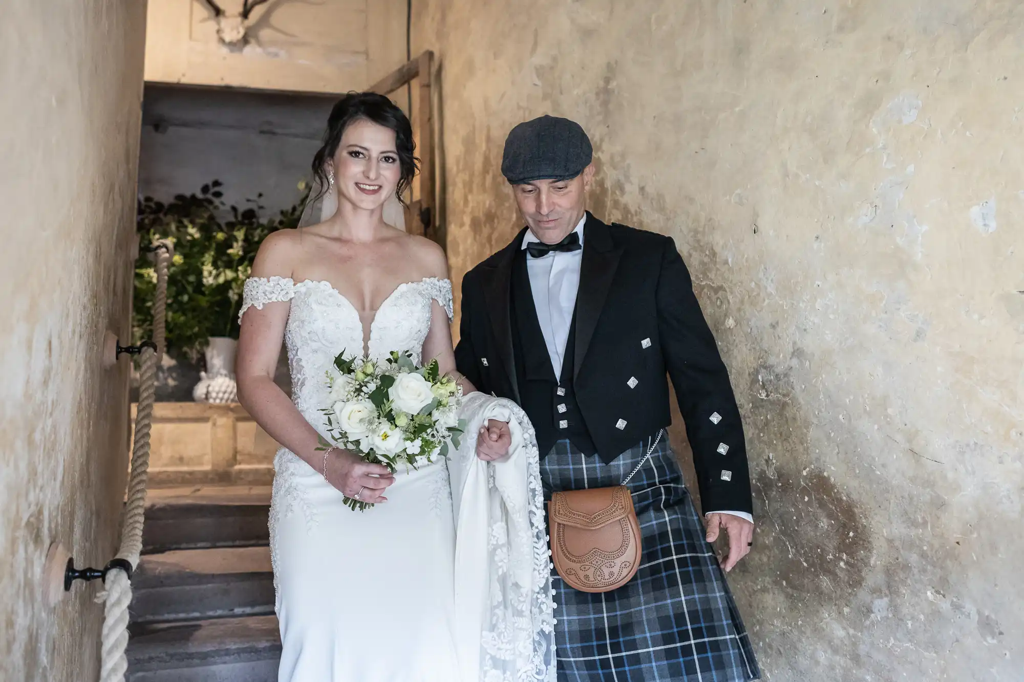 A bride in a white gown holding a bouquet stands next to a man in a traditional Scottish outfit, including a kilt and sporran, on a stone staircase.