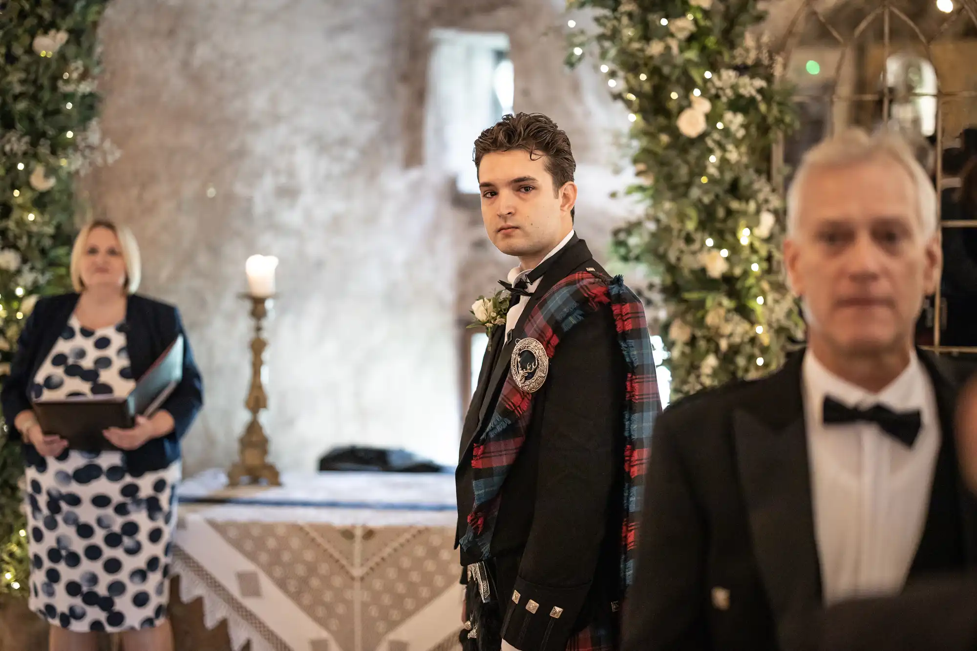 A man wearing a traditional Scottish kilt stands in the center of a decorated room, looking forward. An officiant and another man in formal attire are in the background.