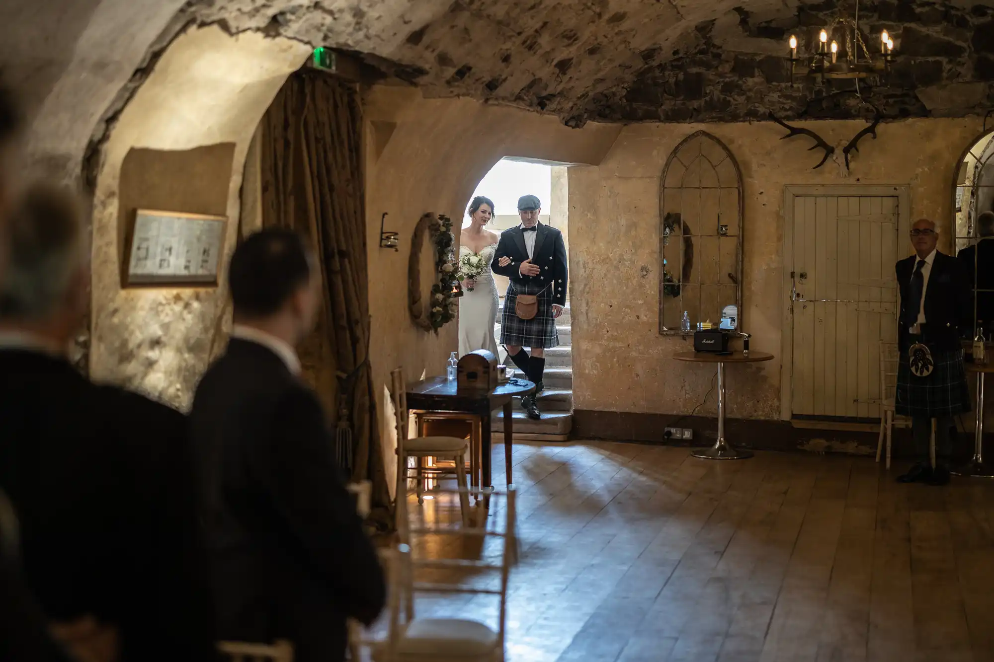 A bride and groom, both in formal attire, walk down an aisle in a dimly lit, rustic stone hall, witnessed by guests.