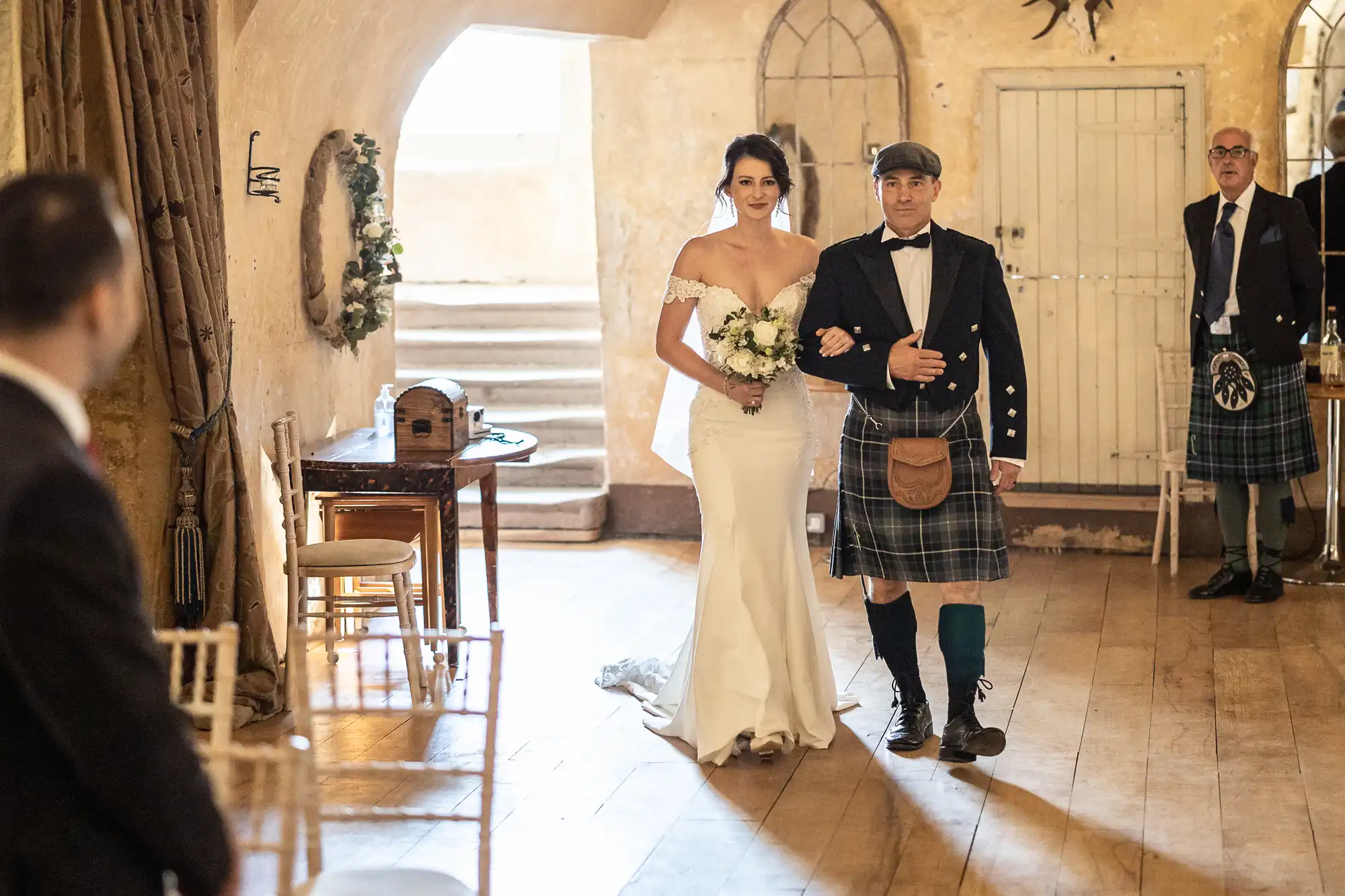 Bride in white gown and bouquet walks down the aisle escorted by a man in a kilt, while another man in a kilt stands behind them inside a warmly lit room.