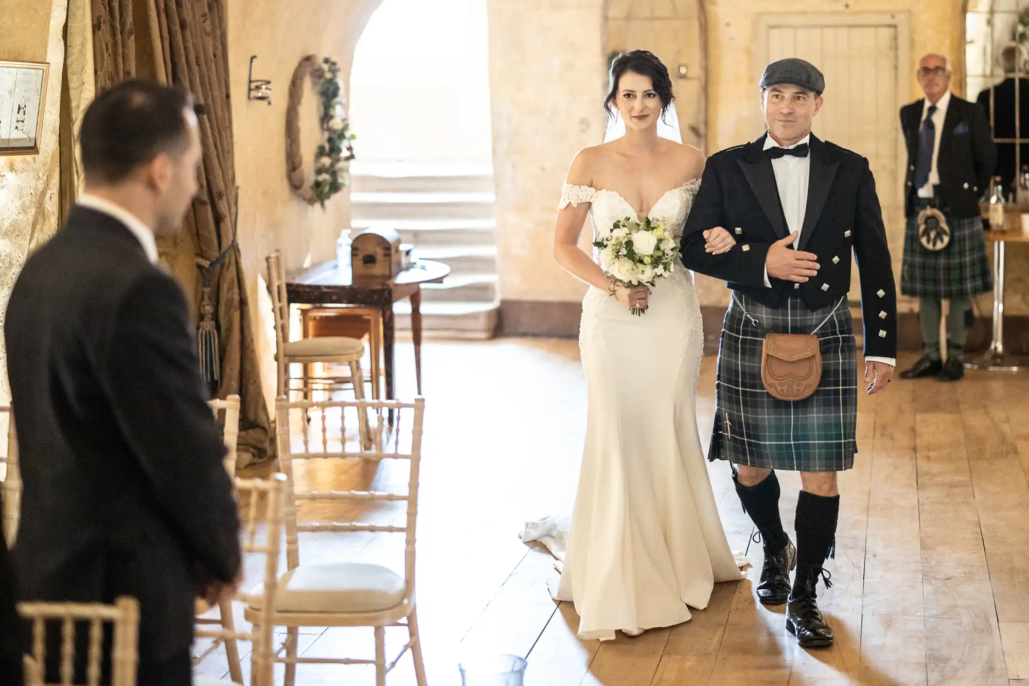 A bride in a white dress holding a bouquet walks down the aisle on the arm of a man in a kilt, while a person in a suit stands at the front and another man in a kilt stands nearby.