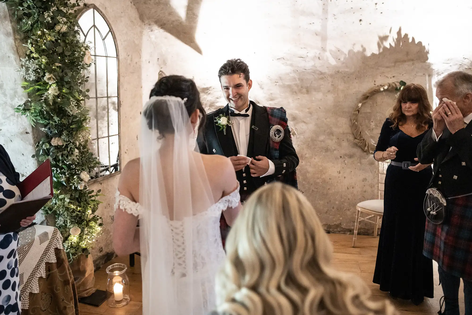 A bride and groom face each other during their wedding ceremony in a dimly lit room with rustic decor. The groom is smiling and dressed in a kilt, while guests stand around observing the moment.